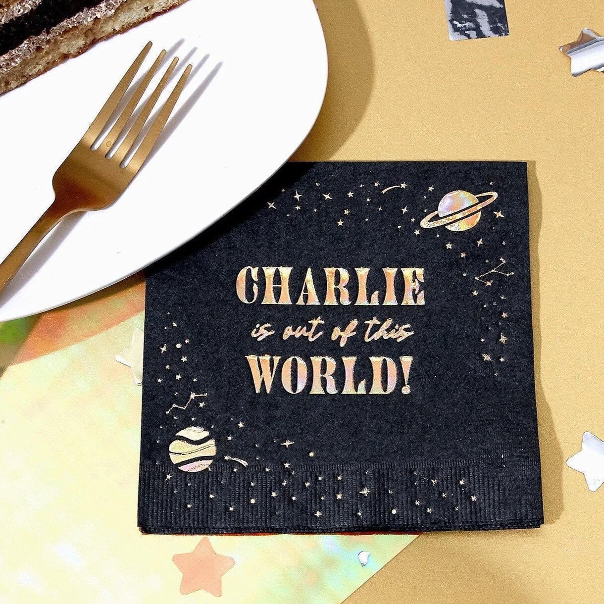 A black party napkin with space-themed illustrations and the quote "Charlie is out of this world!" placed on a table near a white plate and a gold fork, with some star-shaped and other decorative metallic confetti around.