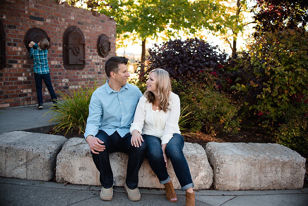  husband and wife sit by a brick wall while their son plays peek a boo behind them 