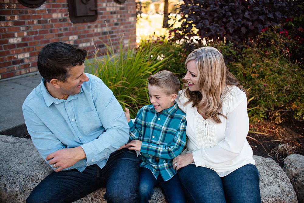  mom and dad sit by a brick wall in Bend, Oregon while tickling their son 