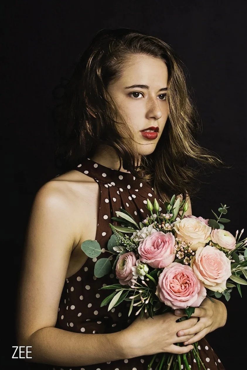 A young woman with shoulder-length brown hair holding a bouquet of pink and white roses and greenery wearing a brown polka-dot sleeveless dress against a black background