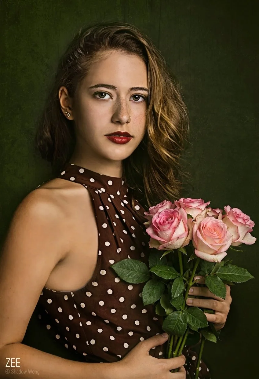 A young woman with shoulder-length wavy brown hair and freckles, wearing a brown sleeveless polka dot dress, holding a bouquet of pink roses, standing against a dark green background.