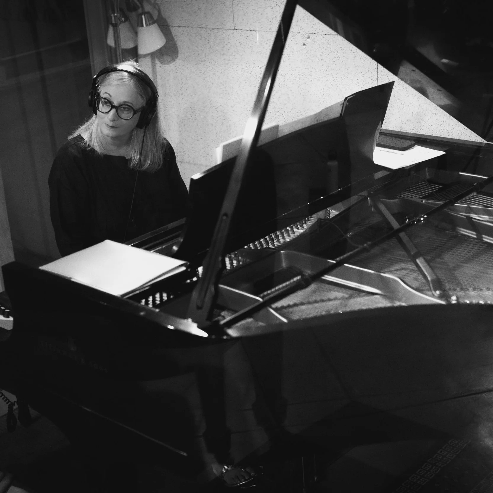 A woman wearing glasses and headphones sits at a grand piano in a recording studio. Renee Rosnes, Piano