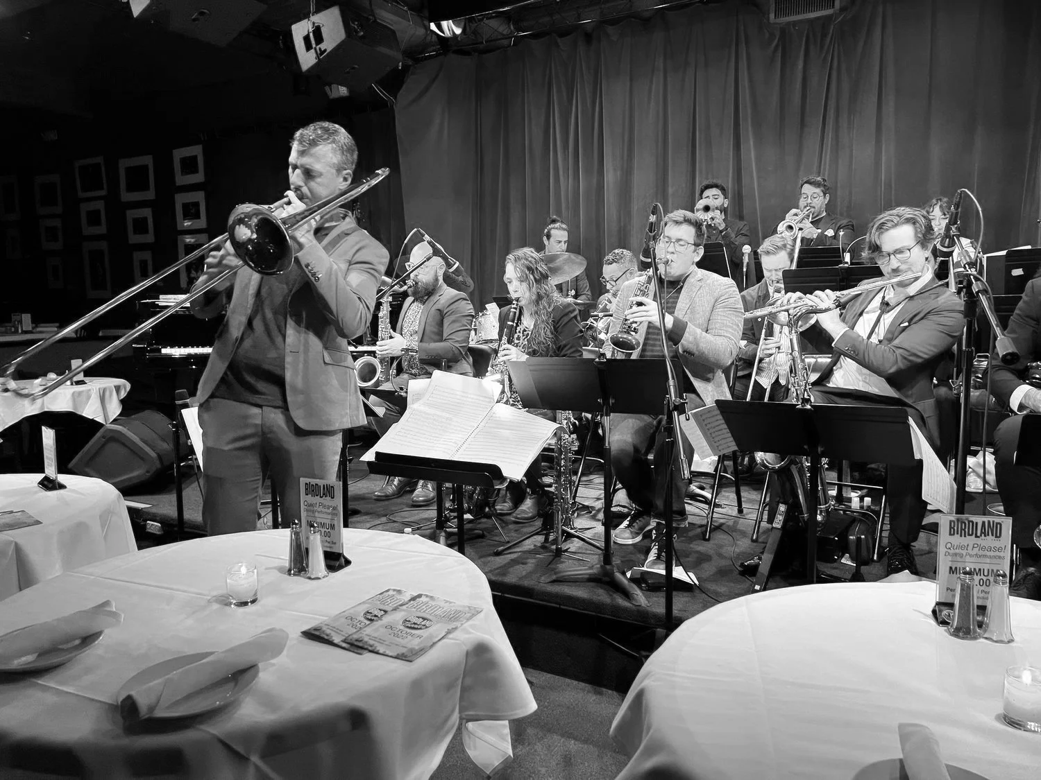 A jazz band performing on stage with multiple musicians playing instruments such as saxophones, trumpet, trombone, and piano. The band members are dressed in formal attire, and in the foreground, there are empty tables with white tablecloths, napkins, and menus or flyers on top
