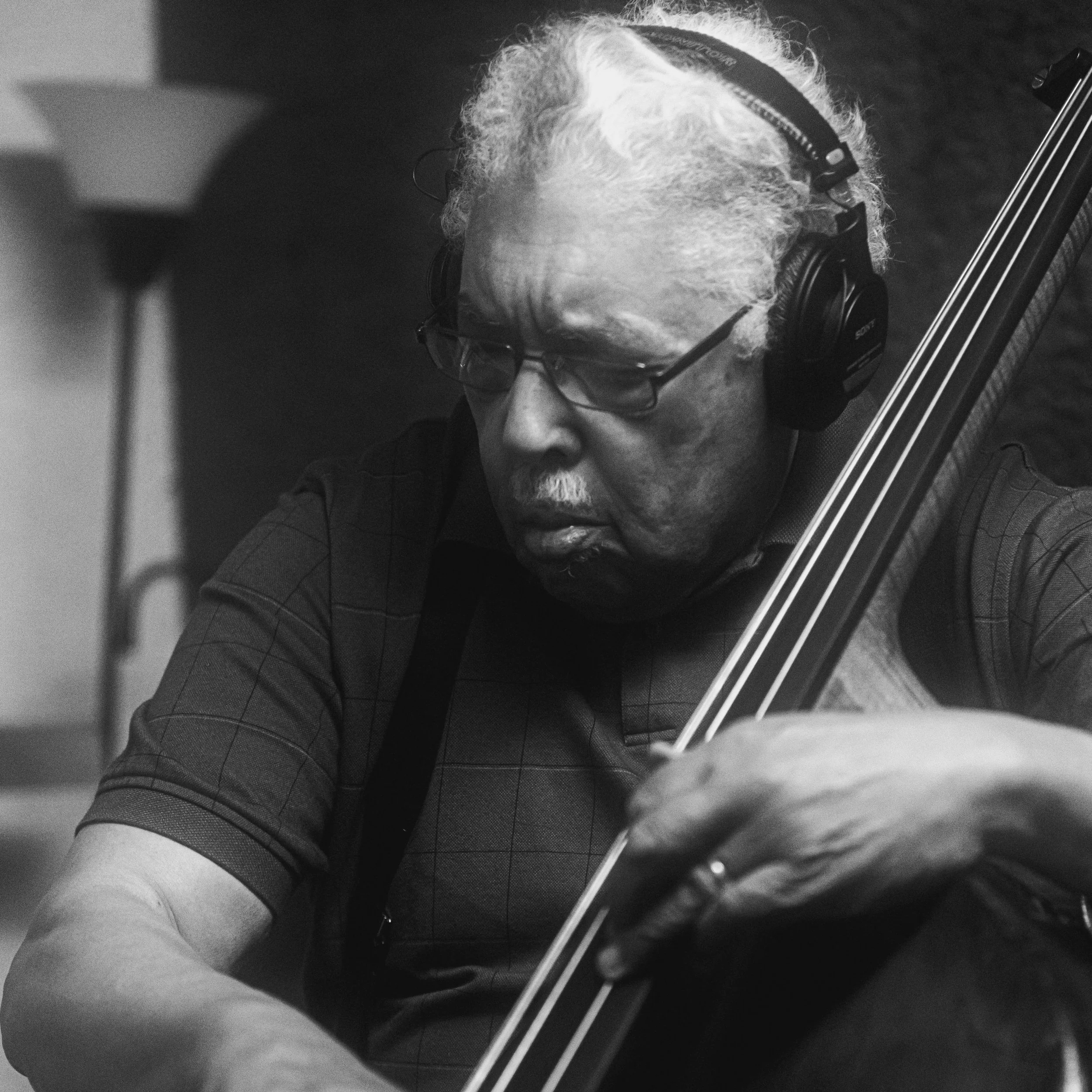 An elderly man with curly hair, glasses, and wearing headphones playing a string instrument. This is Rufus Reid