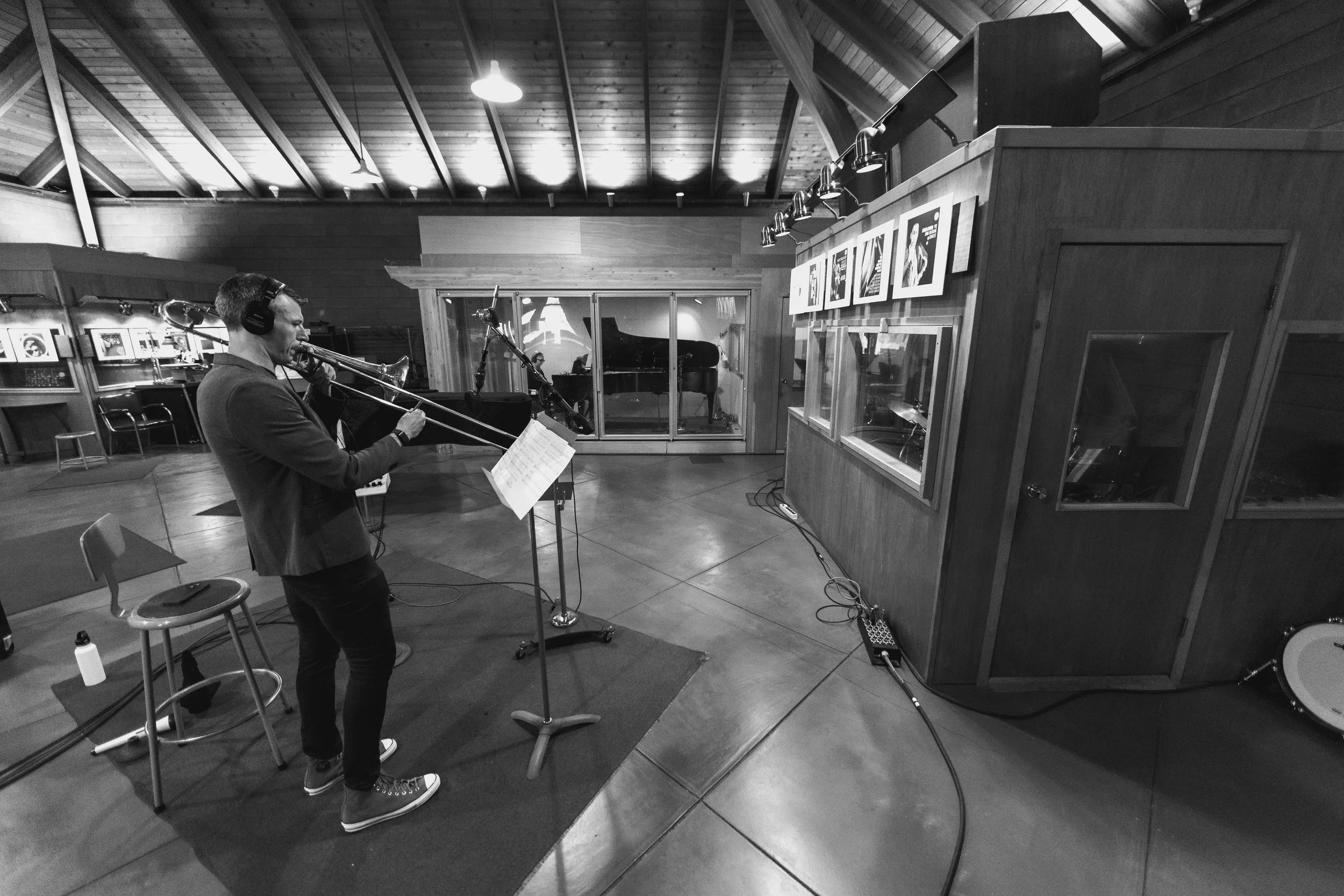 A man wearing headphones playing trombone in a recording studio with a grand piano in the background and framed artwork on the walls.