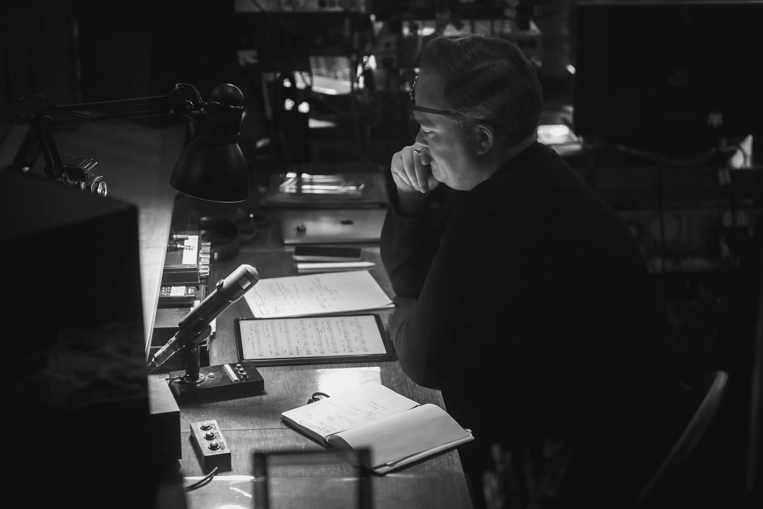 A man with glasses sitting at a desk, looking at a tablet, surrounded by papers and notebooks, in a dimly lit room. Ryan Truesdell, Producer