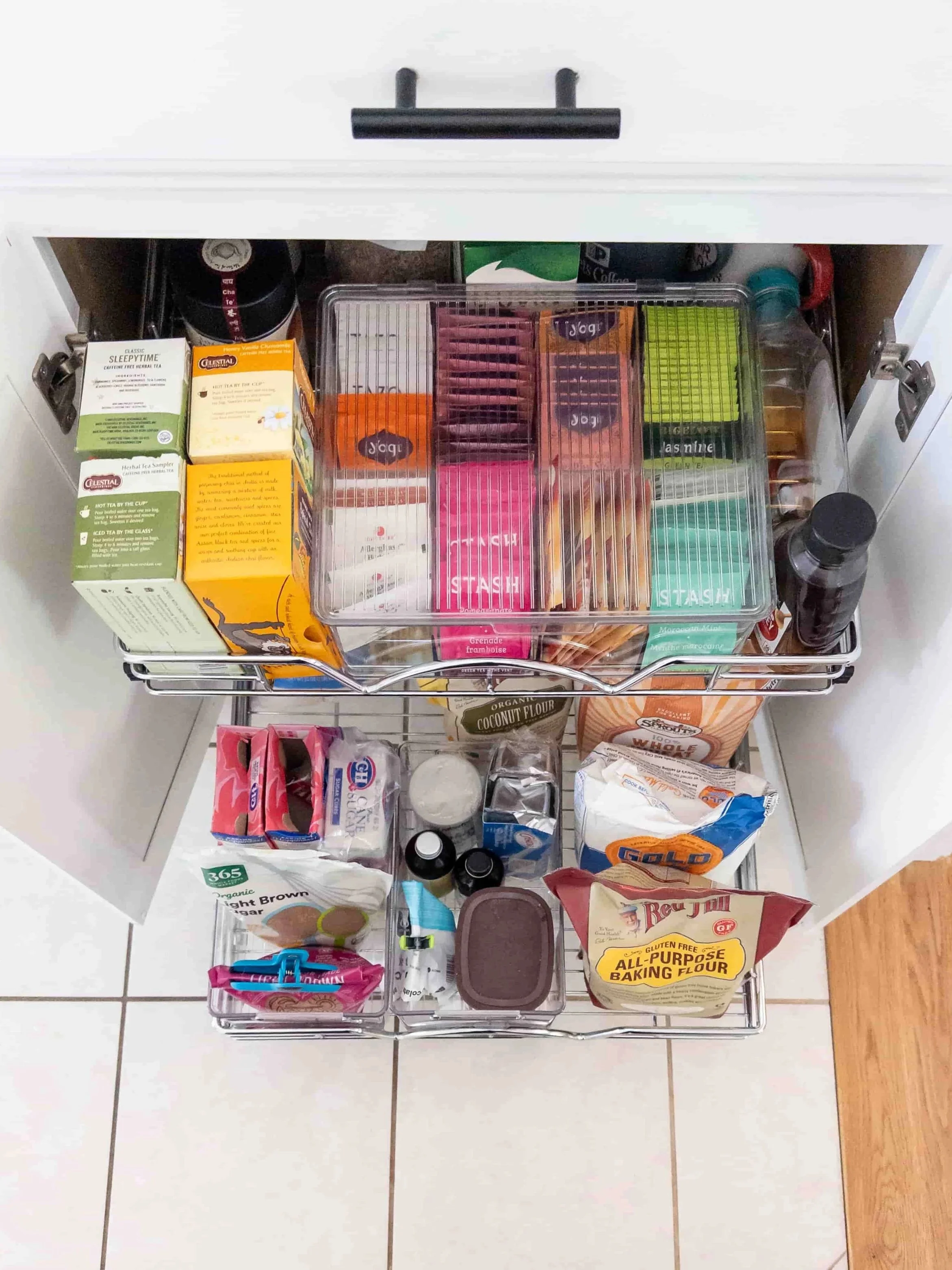 Open kitchen drawer containing various packaged food items including tea, flour, baking powder, sugar, snacks, and other pantry essentials.