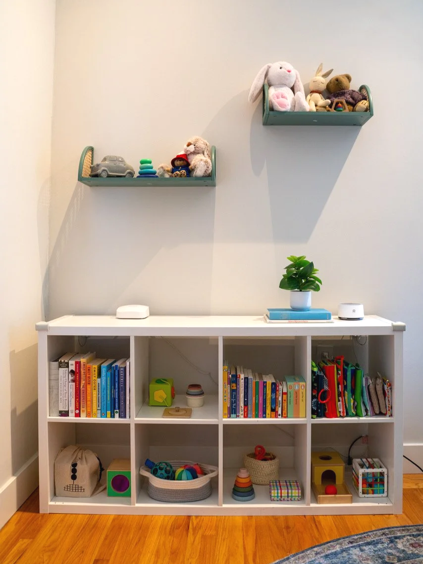 A white cubed bookshelf with children’s books, toys, and storage bins, topped with a potted plant. Above, two floating shelves hold stuffed animals, toys, and a car model. The setting appears to be a child's playroom or reading area.