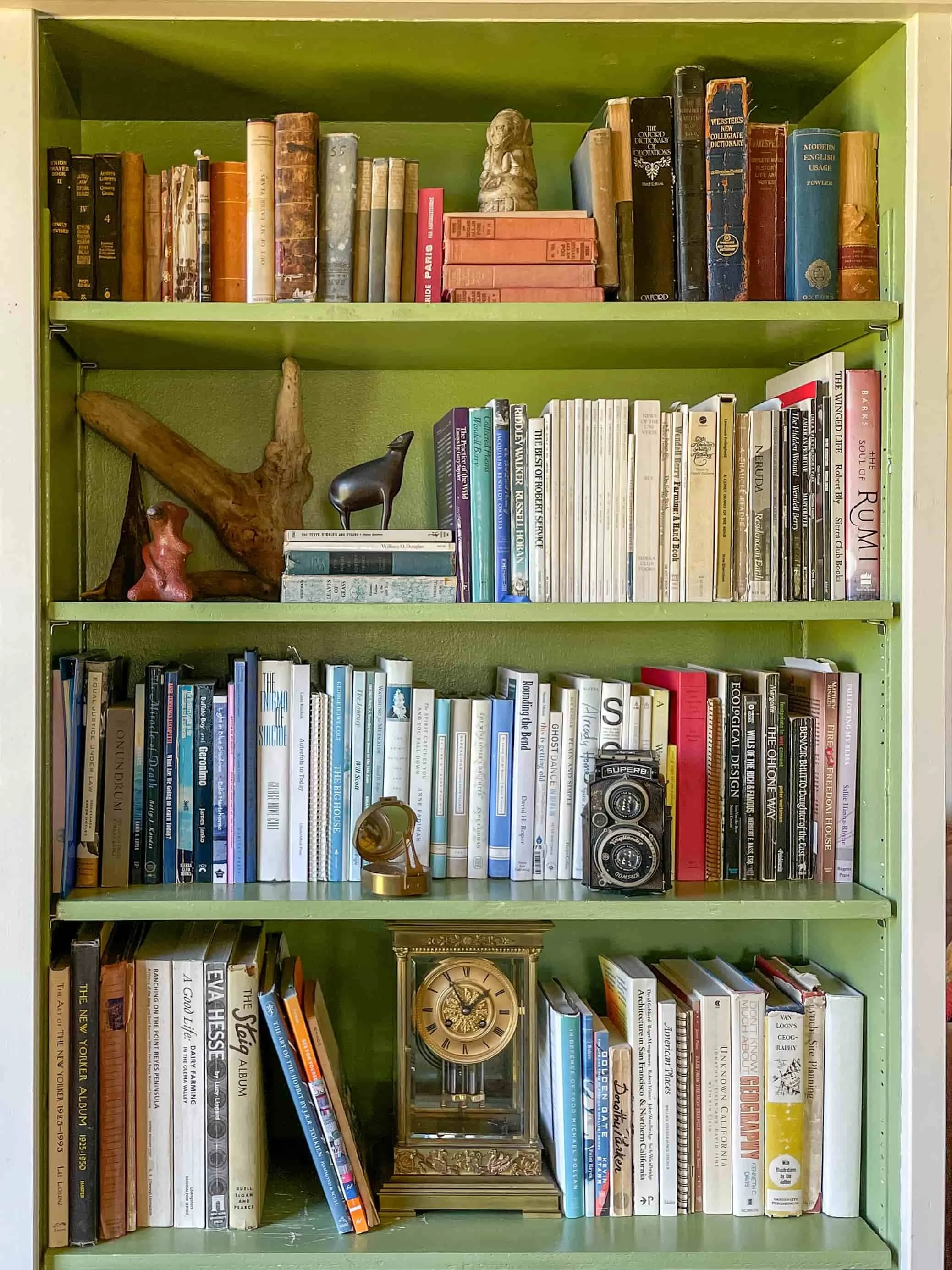 A green bookshelf, with four shelves, is styled with books, an antique glass pendulum mantel clock and a mix of keepsakes.