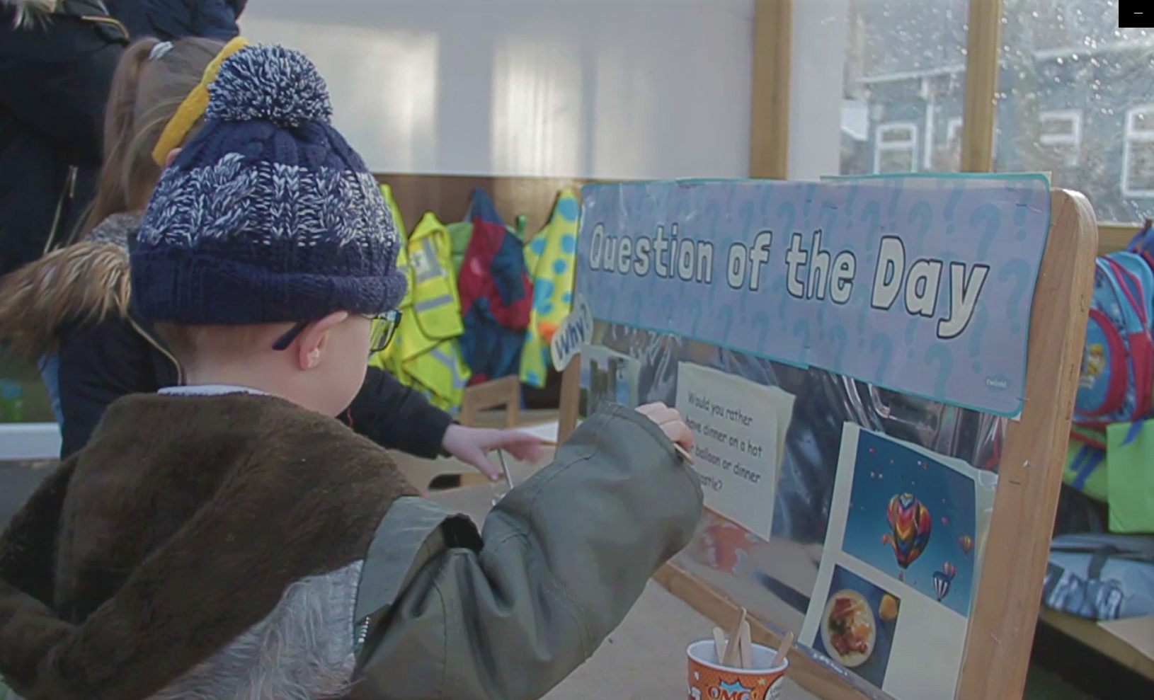 Reception children placing lolly sticks in one of two cups to show whether they’d rather have dinner in a castle or a hot air balloon. The cups are next to a board titled Question of the Day and there are pictures illustrating the two options.