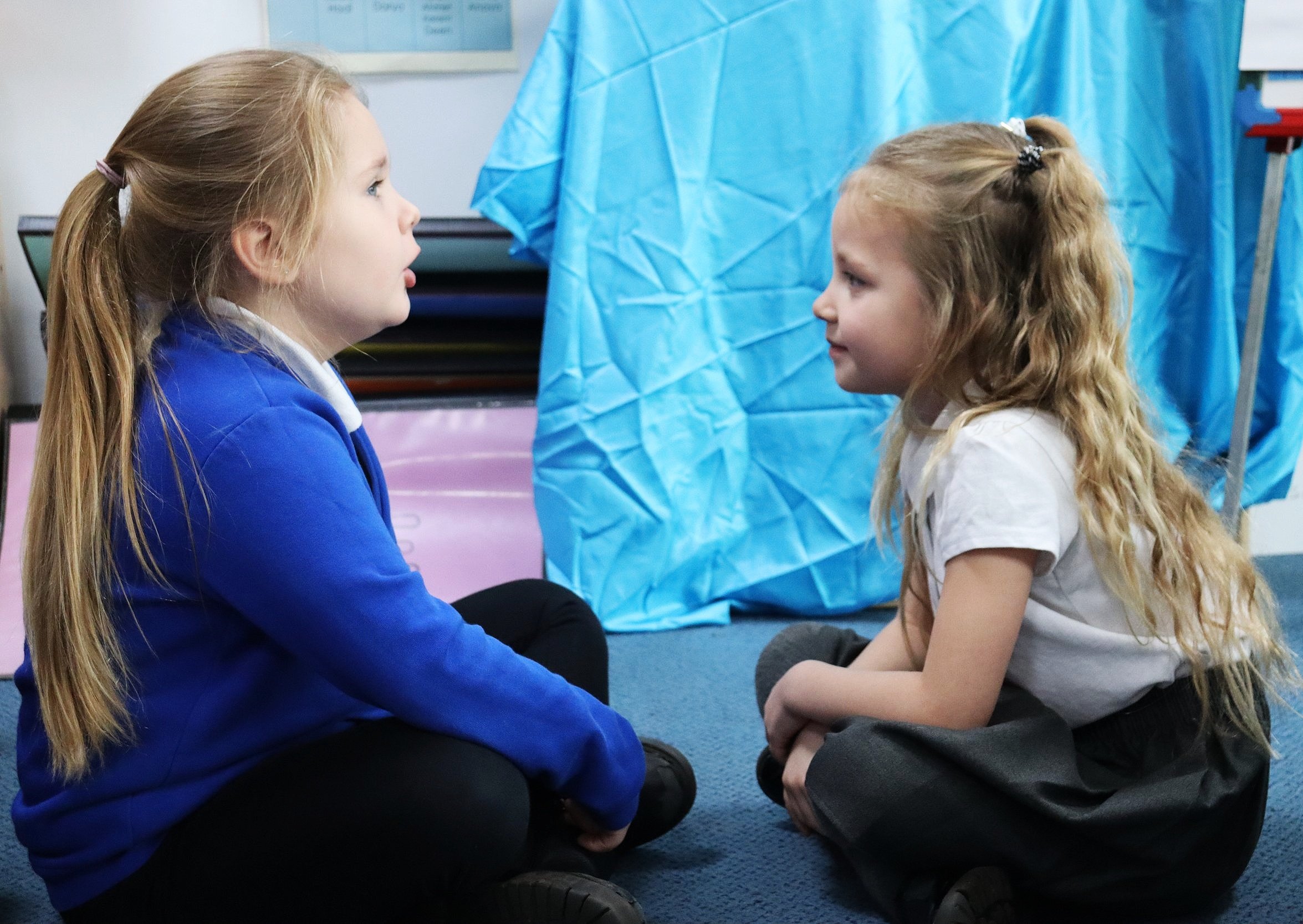A pair of pupils sit cross-legged, one speaking and the other listening closely