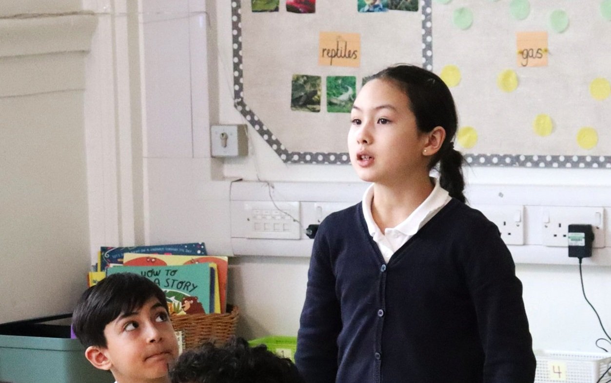A student speaking, standing at her desk, while another gives her attention