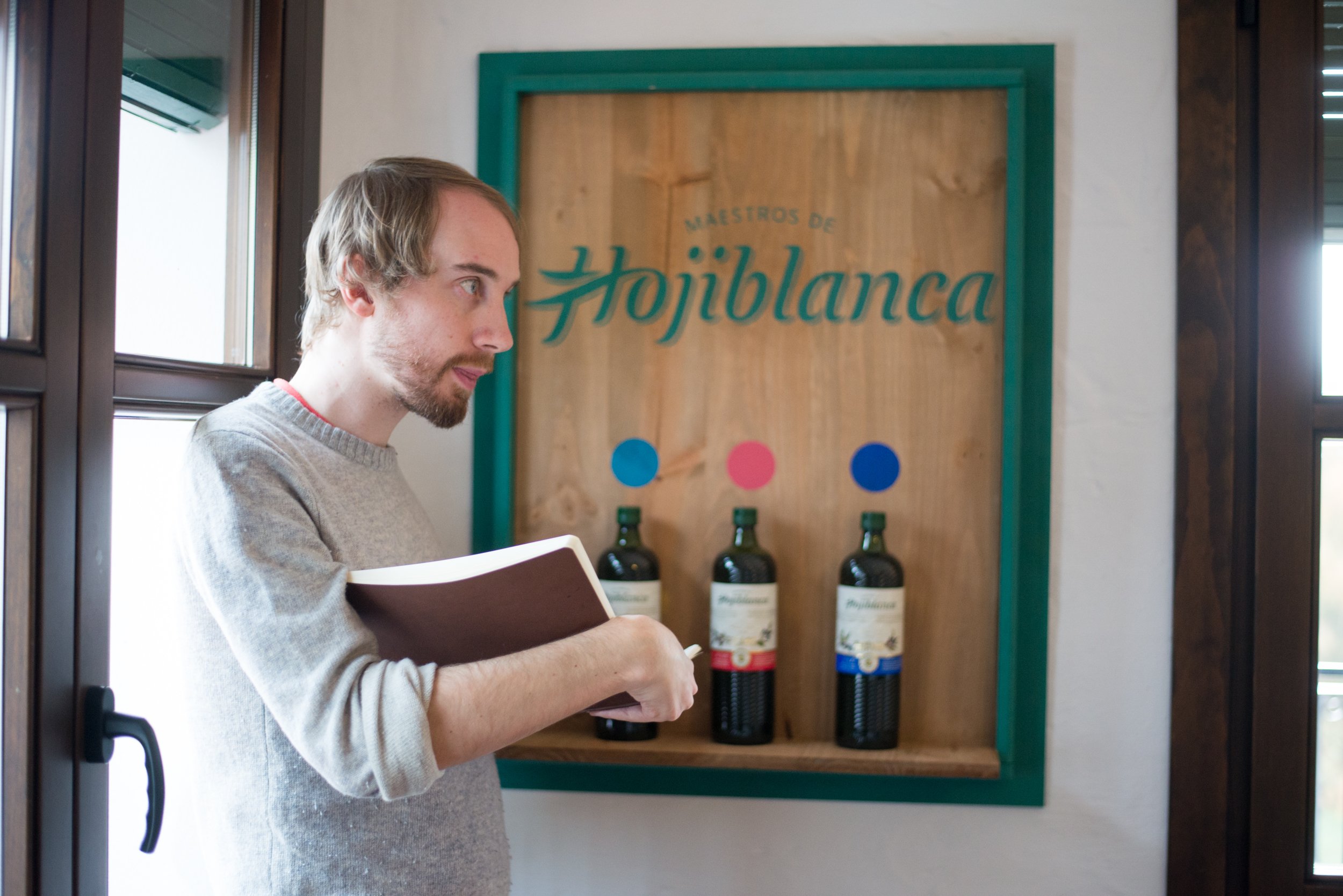 Hombre con libro en mano observando cartel de madera con botellas de vino en una bodega, en el fondo se lee 'Maestros de Hojiblanca'