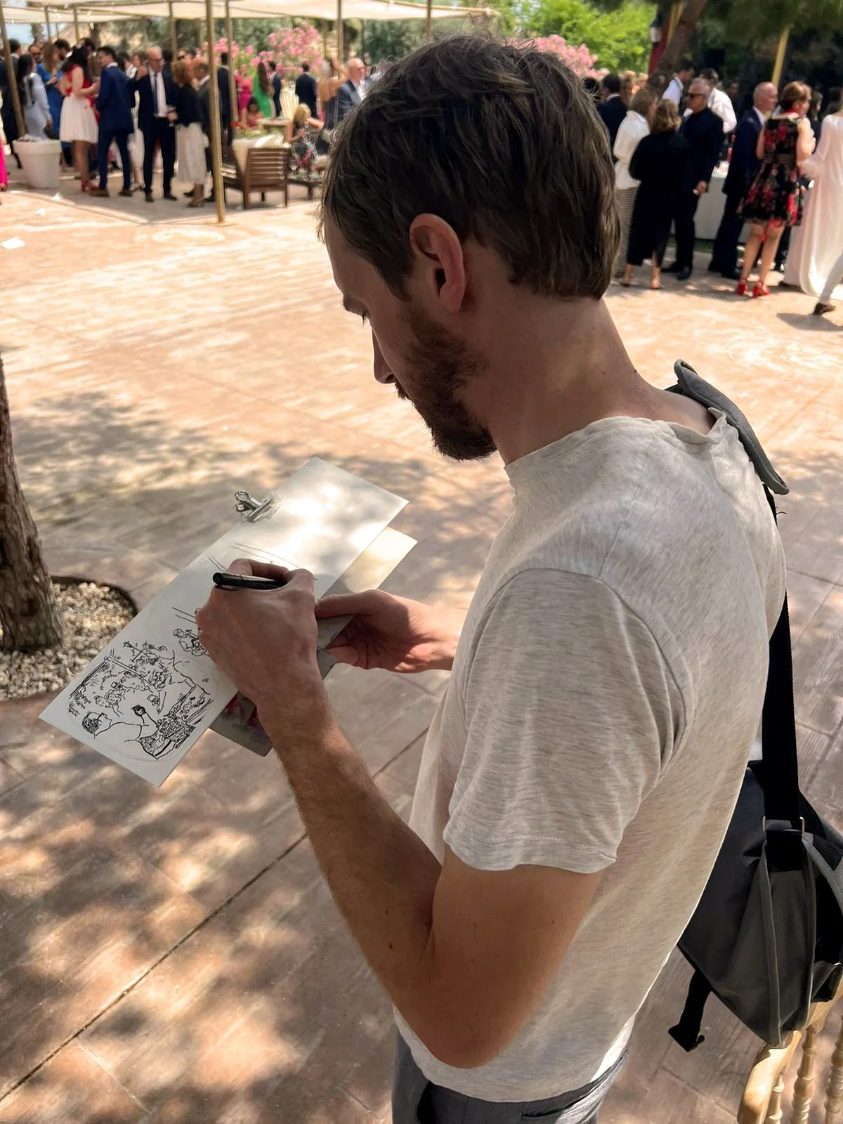 Un hombre firmando un dibujo en un evento al aire libre con muchas personas en el fondo.