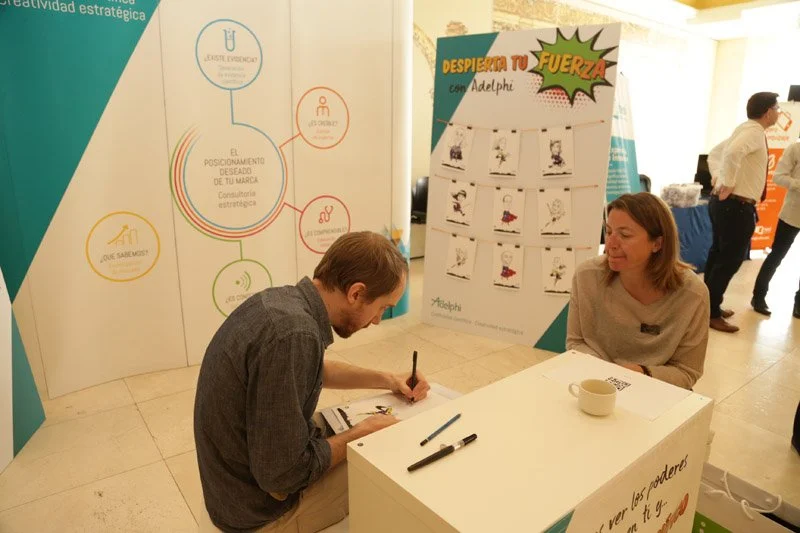 Persona en una mesa firmando un libro o documento, con otra persona sentada al frente observando. Fondo con carteles y posters relacionados con motivación y estrategia de marca en un entorno de evento o feria.