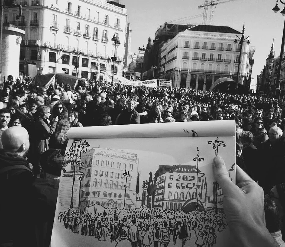Una multitud de personas en una manifestación en una ciudad, con edificios históricos y árboles en el fondo, mientras alguien sostiene un cuaderno con un dibujo de la escena.
