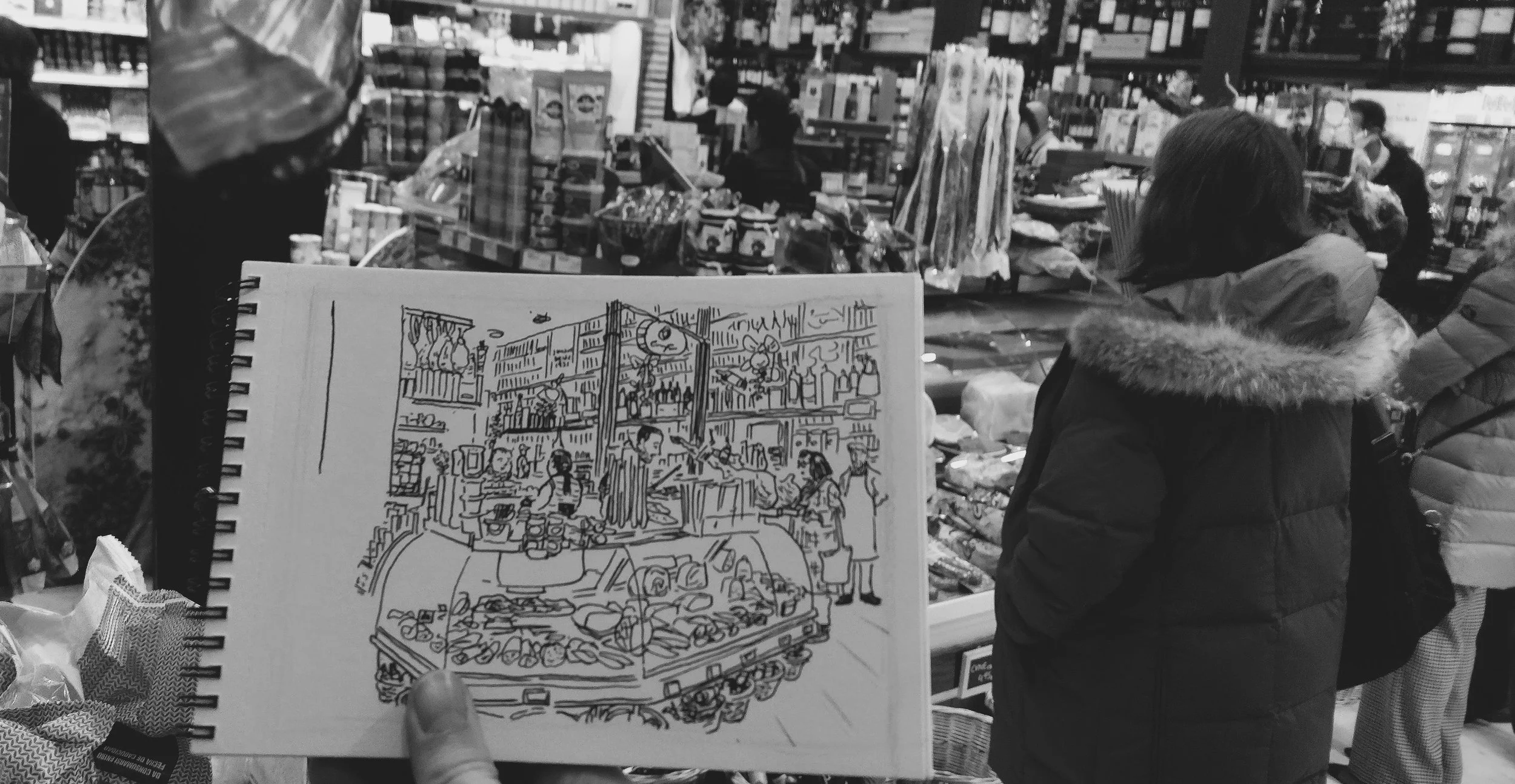 Una persona sostiene un cuaderno con un dibujo de un mercado en una tienda de comestibles, con estantes llenos de productos y varias personas comprando.