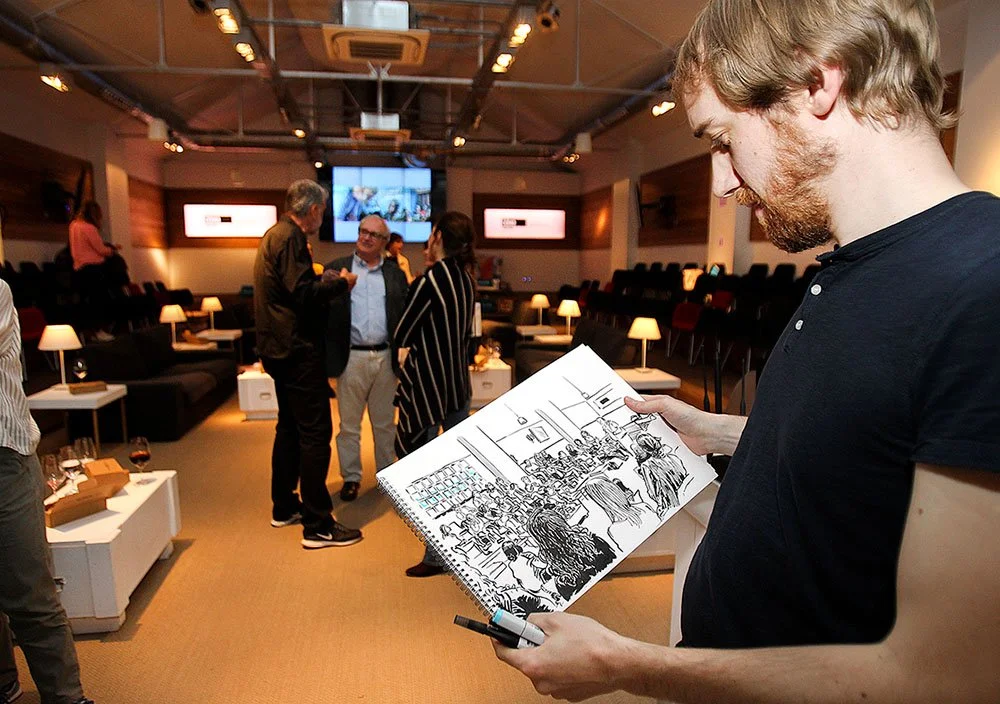 Un grupo de personas conversando en una sala de reuniones o evento, con una persona en primer plano que sostiene un cuaderno con un dibujo de un evento social.