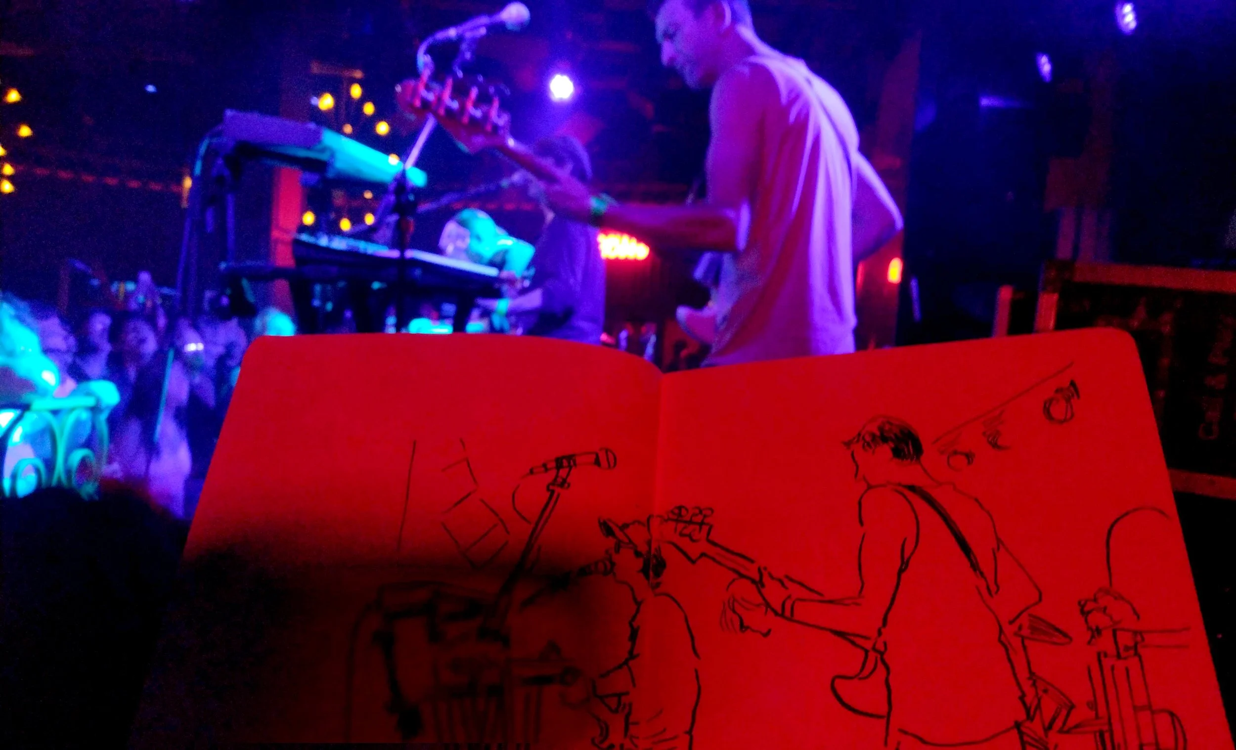 Un grupo de músicos tocando en un concierto oscuro con luces violetas y azules, con una multitud admirando en el fondo; en primer plano, un cuaderno rojo con dibujos de músicos tocando instrumentos, incluyendo una guitarra y una batería.