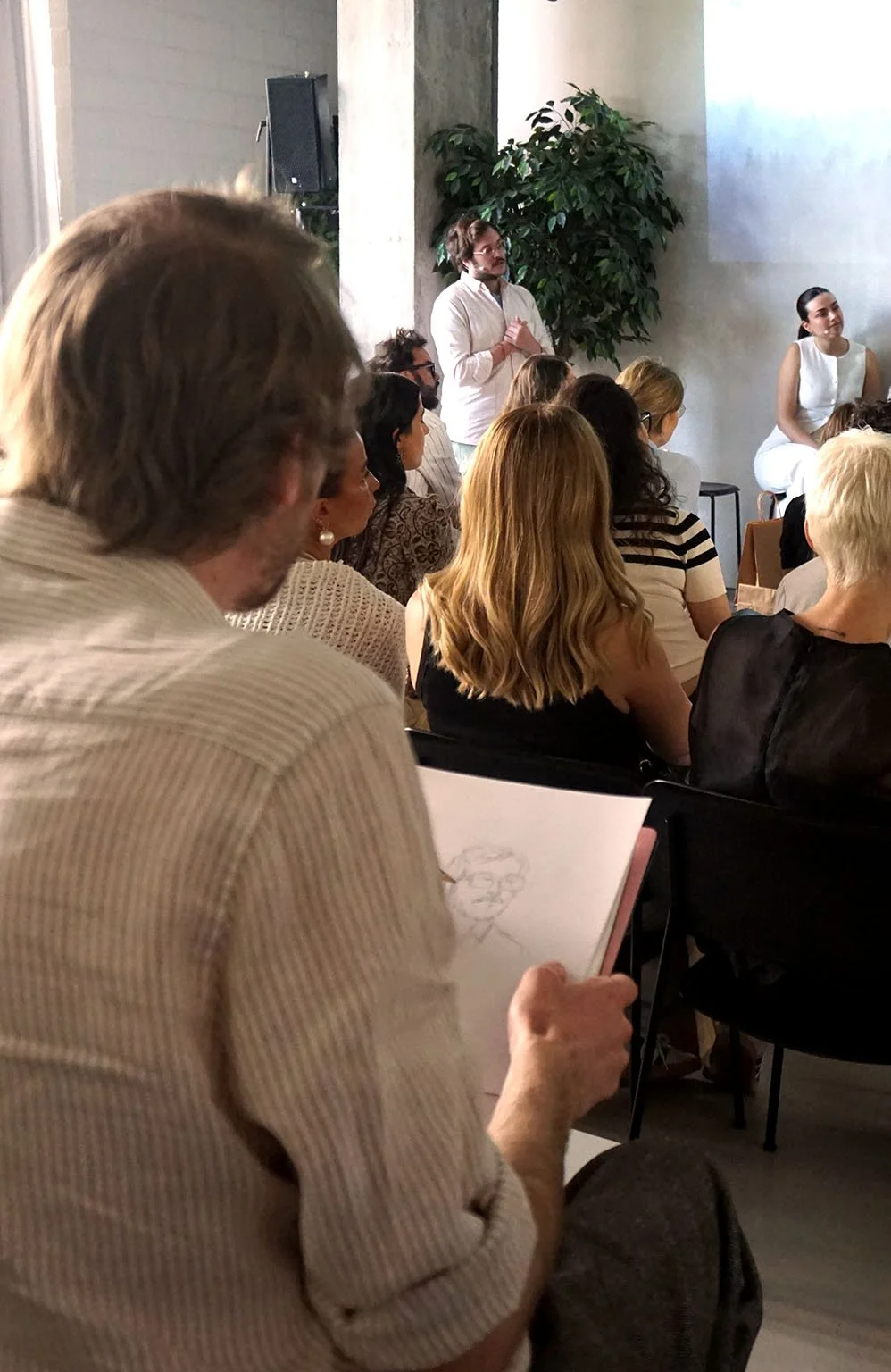 Hombre con barba y gafas en una chaqueta blanca hablando frente a un grupo de personas. Muchas de ellas están sentadas y algunas de ellas tomando notas. Al fondo hay una planta grande y un muro de cemento.