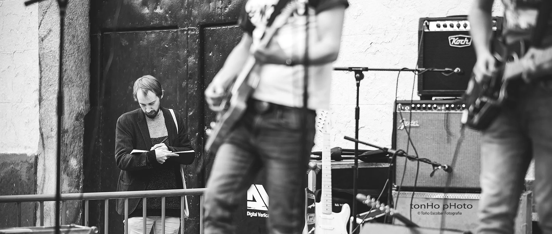 Un hombre con barba y chaqueta observa su teléfono móvil en un entorno de concierto callejero, con instrumentos musicales y amplificadores a su alrededor.