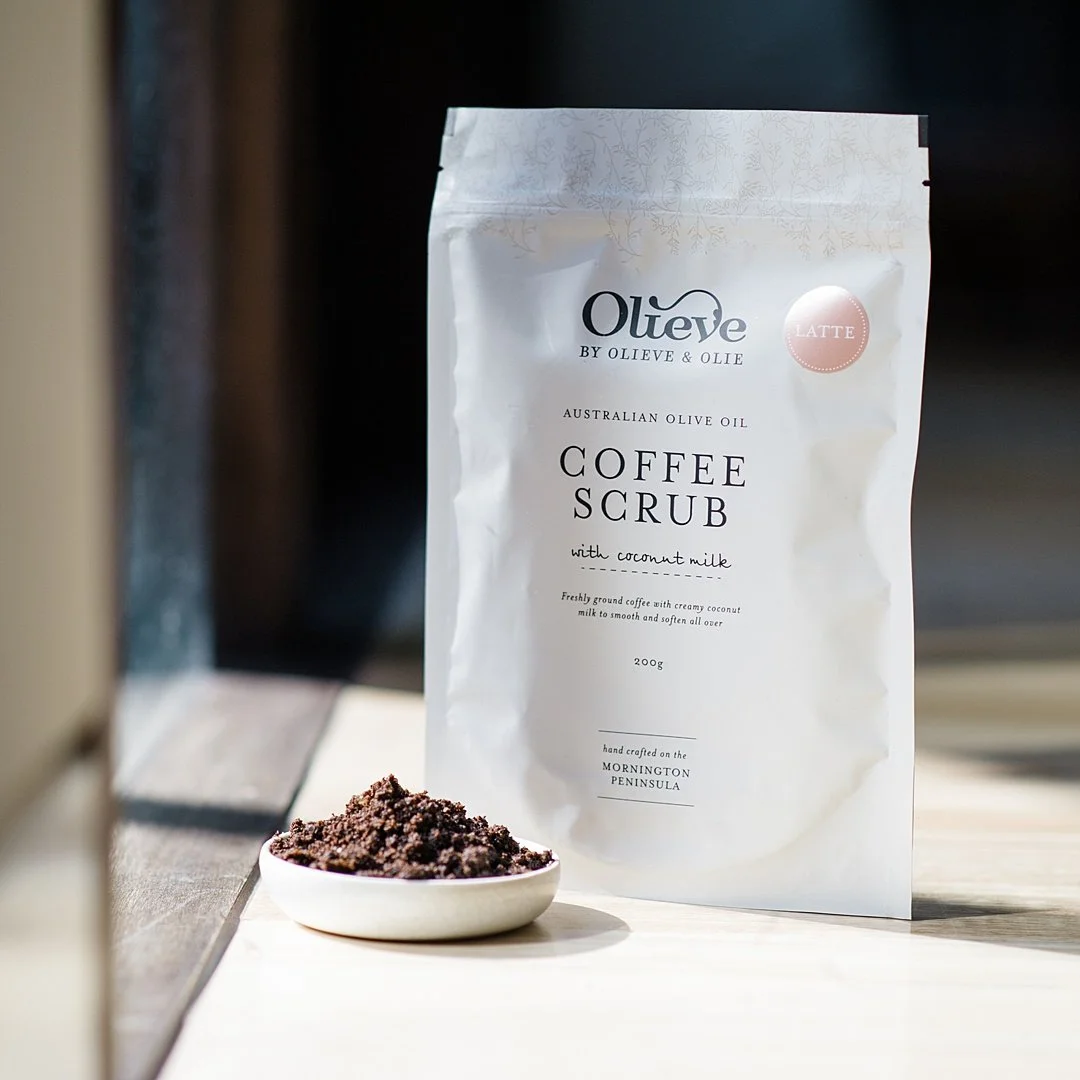 pouch of coffee scrub on a table near sunlight next to a small white bowl of coffee grounds body scrub