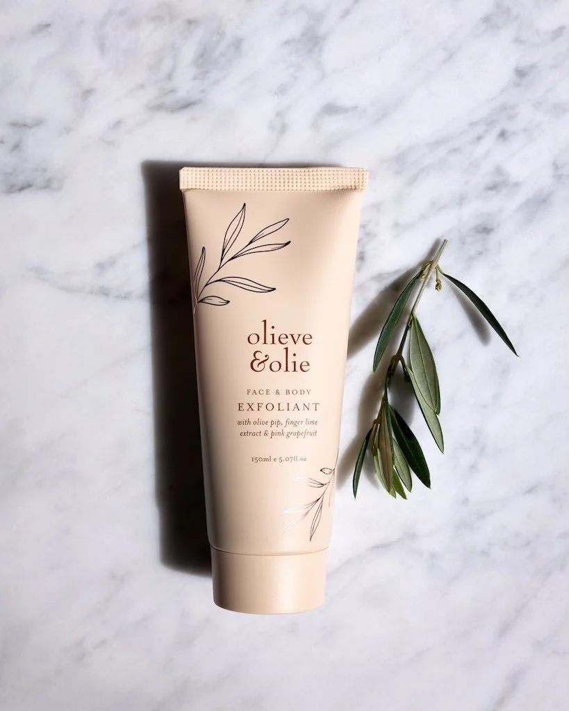 Beige tube of Face & Body Exfoliant on marble backdrop with twig of olive branch.