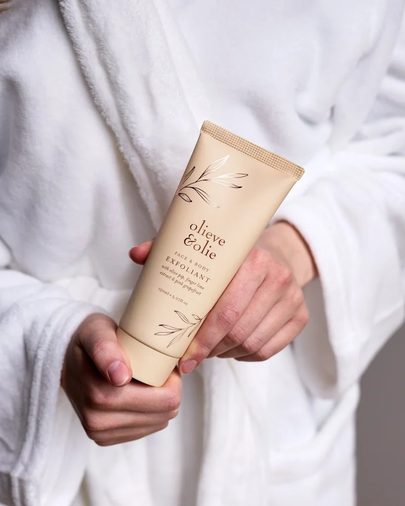 person in white dressing gown holding a beige tube of Face & Body Exfoliant