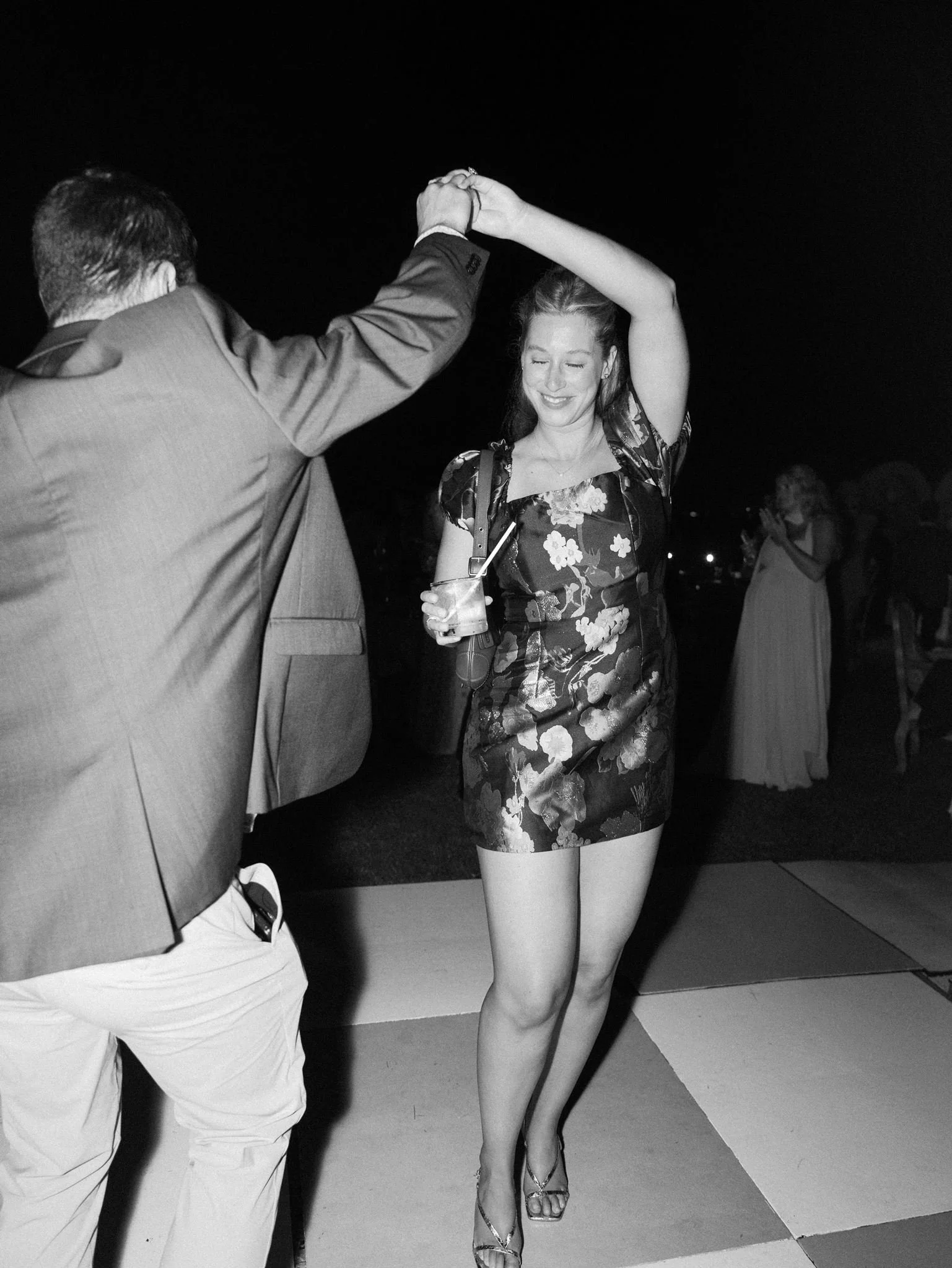 A woman in a floral dress is dancing with a man in a suit at a social event, holding drinks, at night.
