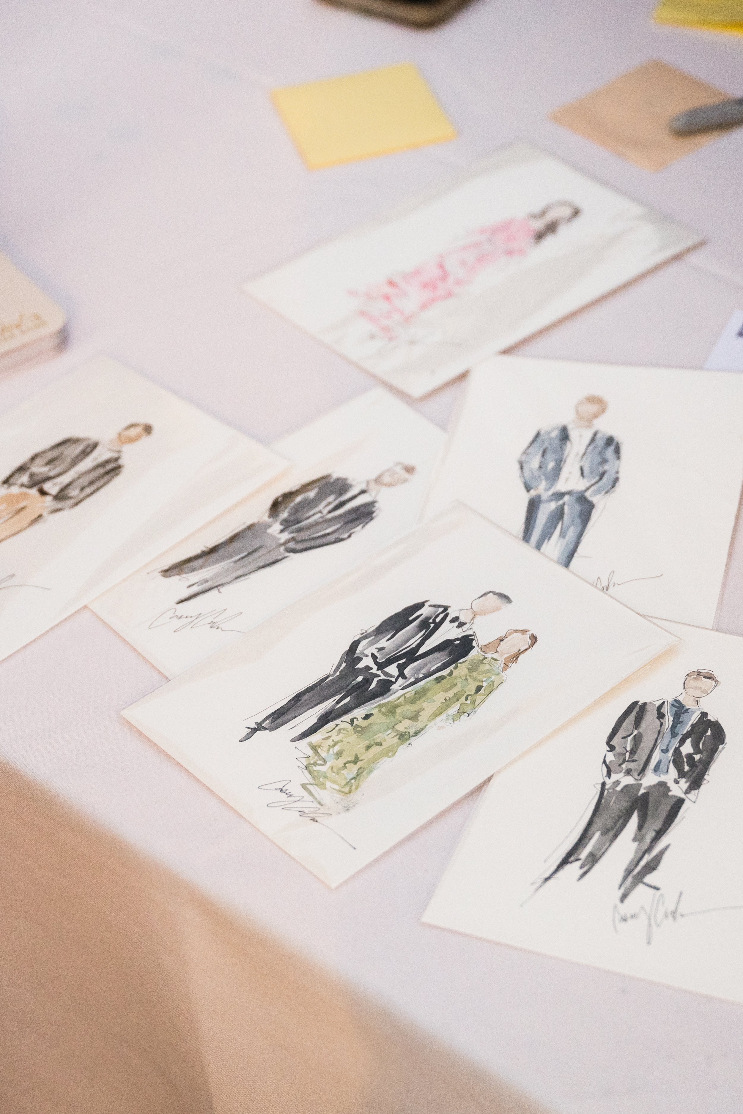 Collection of watercolor portraits of men and women, some standing and some walking, on a pale pink table.