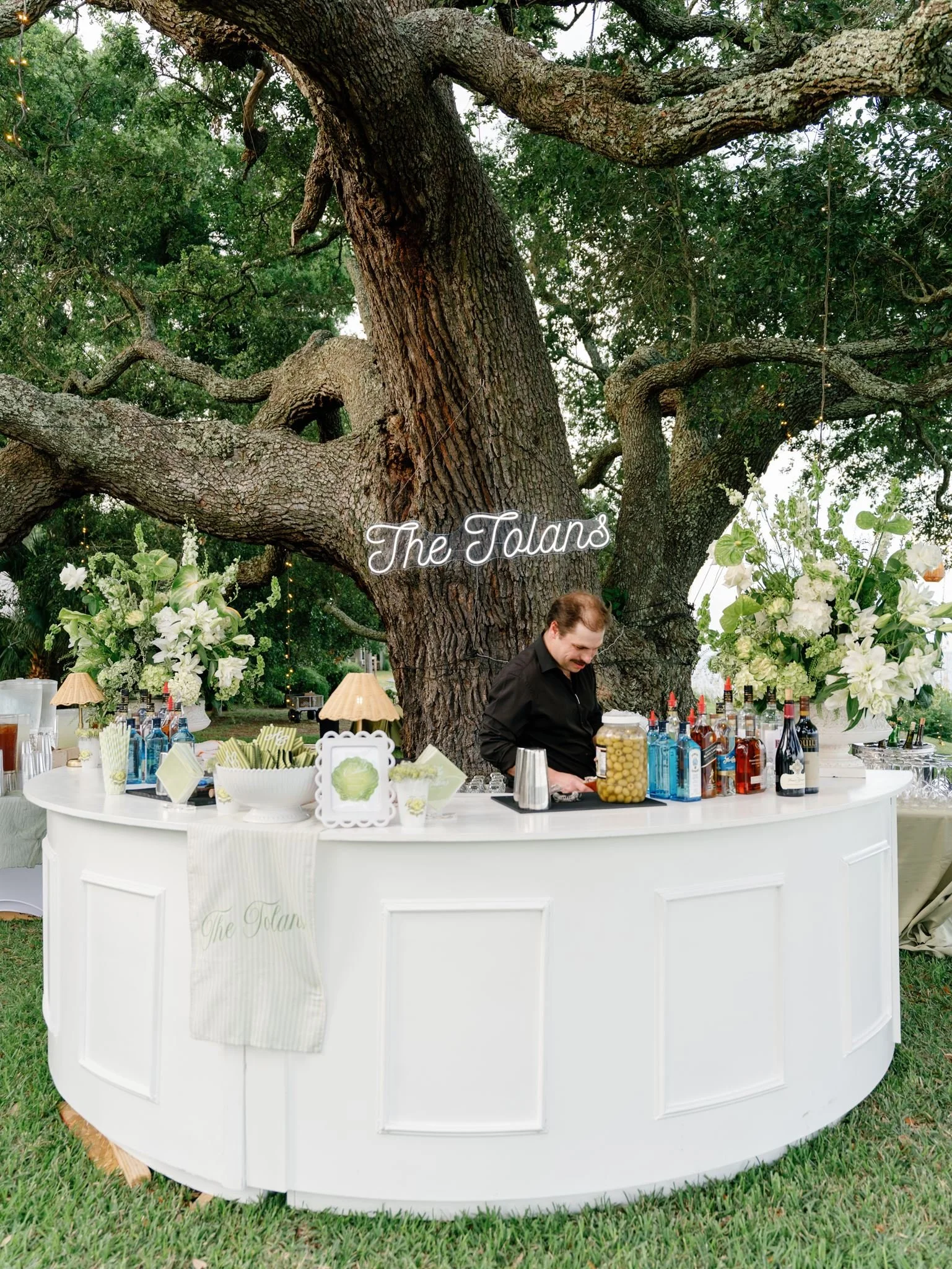 A large tree with thick branches serving as a backdrop for a white bar setup at an outdoor event. The bar is decorated with floral arrangements and has various bottles and bar accessories on top. A man is standing behind the bar. A sign on the tree r