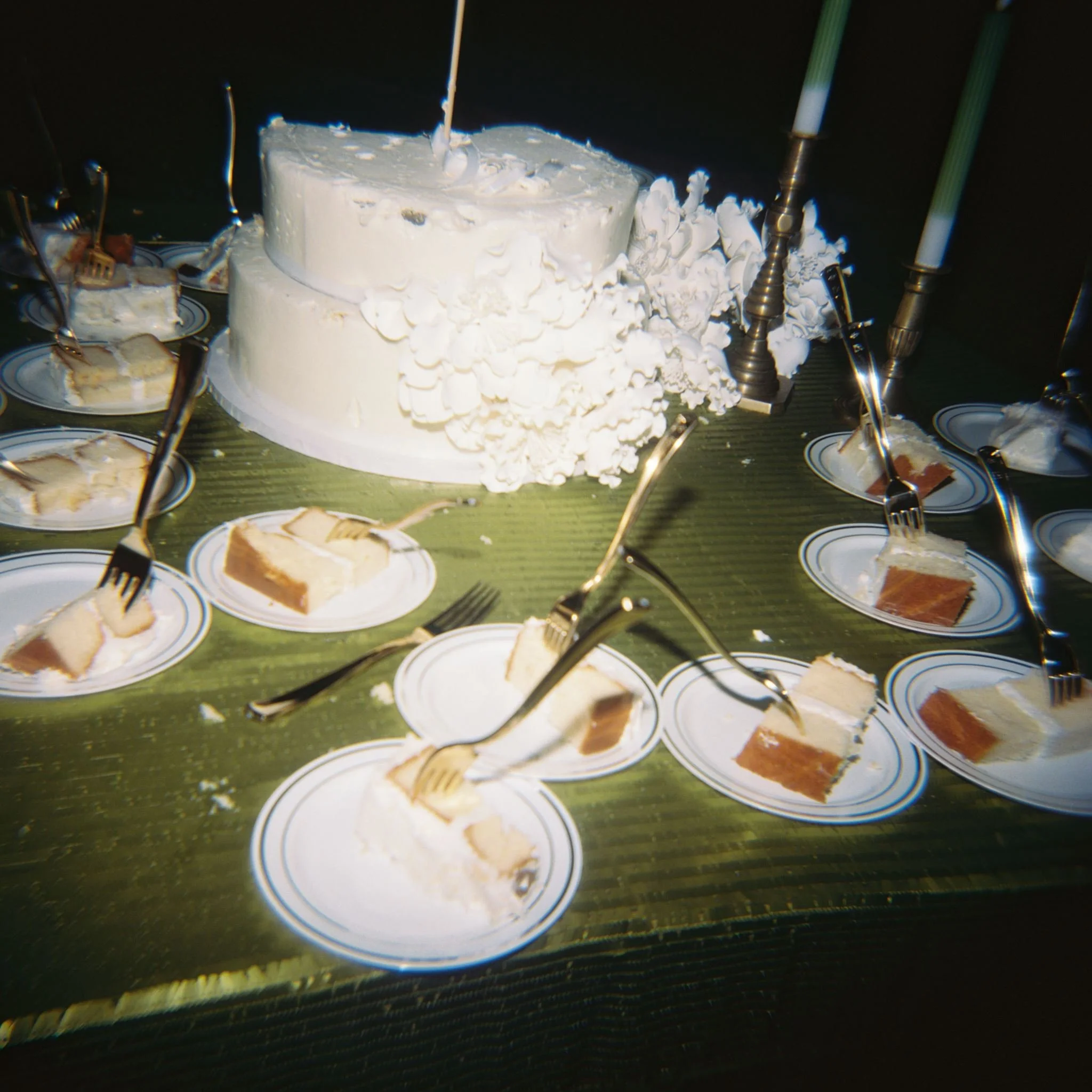 A celebration table with a white frosted cake decorated with white icing, surrounded by plates with slices of cake and forks, and lit candles on the cake.