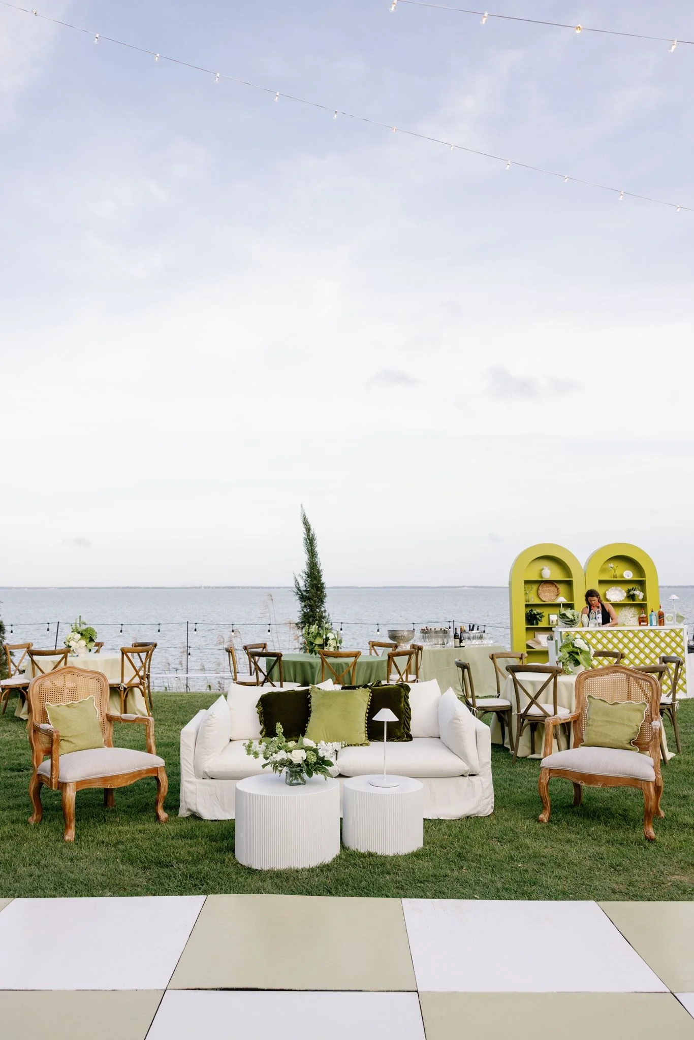 Outdoor event setup near water with floral arrangements, greenery, vintage style chairs, and a white sofa with throw pillows, large yellow shelving unit with decor, string lights in the sky, overcast weather, and a tree in the background.