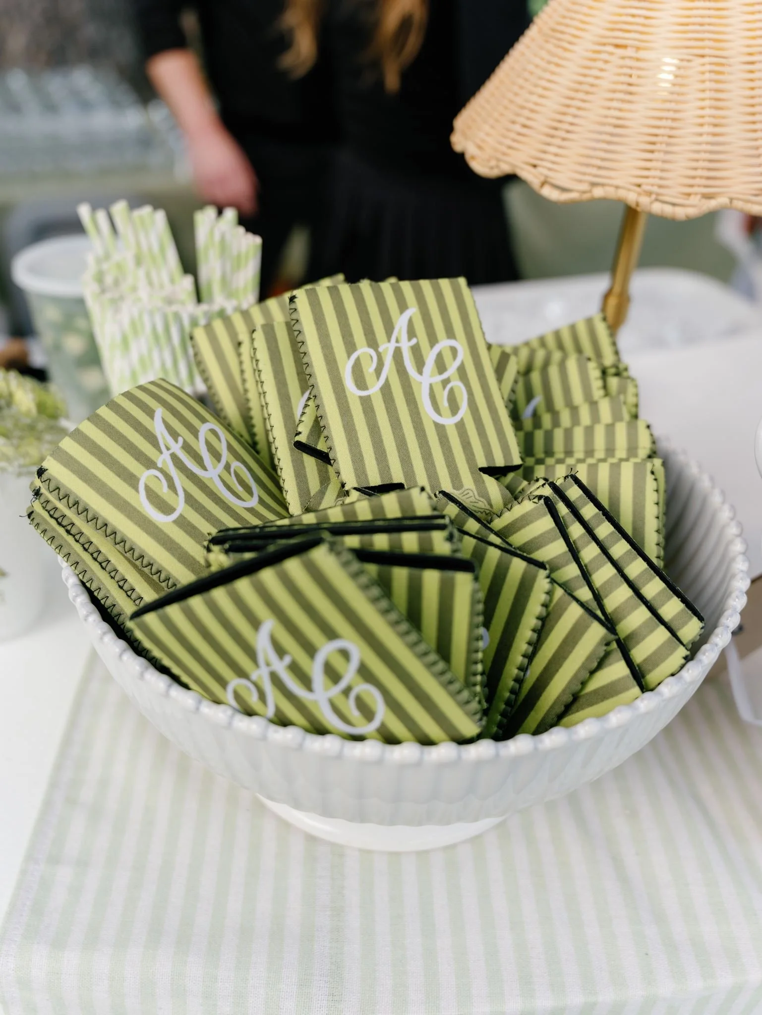 A white bowl filled with green and white striped paper cards with white cursive initials 'A C' on a green striped background, placed on a left side, with a wicker lamp part in the background.