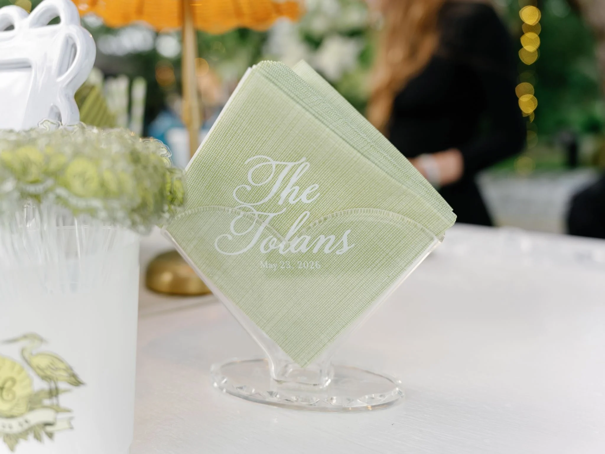 Close-up of a green paper napkin with white cursive text that reads 'The Folsom', standing upright on a clear glass stand, with blurred background that appears to be an outdoor event or market.