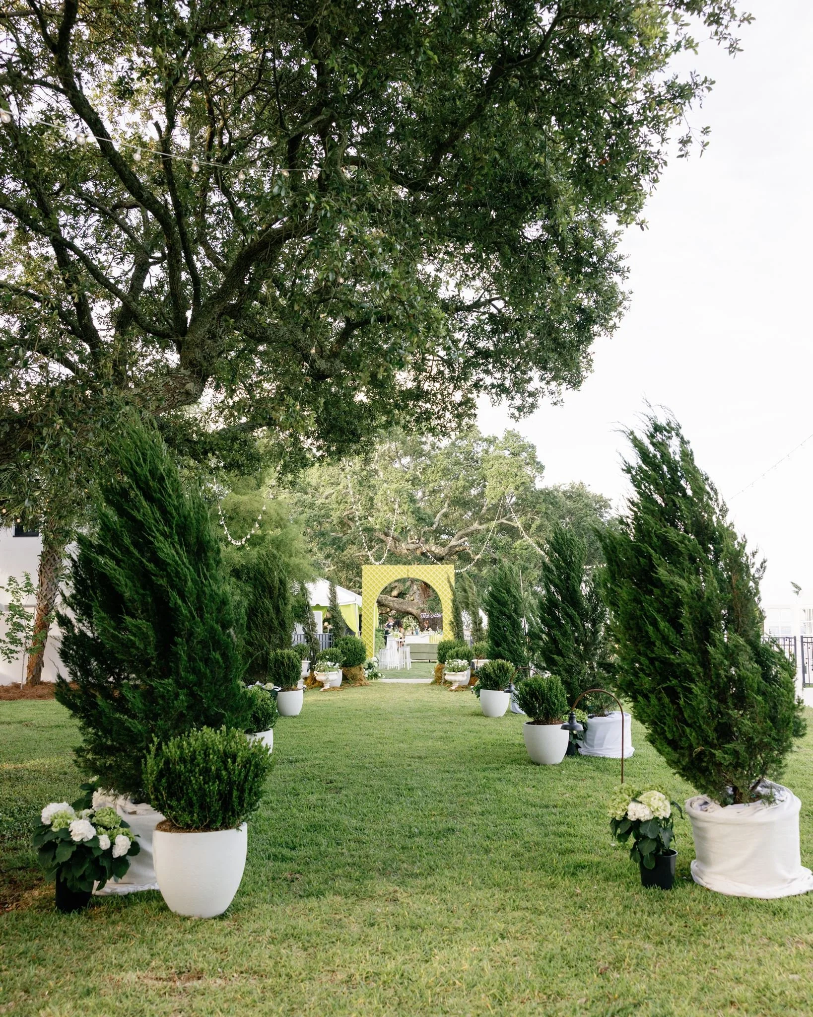 A well-manicured garden setup for an outdoor wedding or event, with white potted floral arrangements, lush green trees, and a decorative yellow archway in the distance.