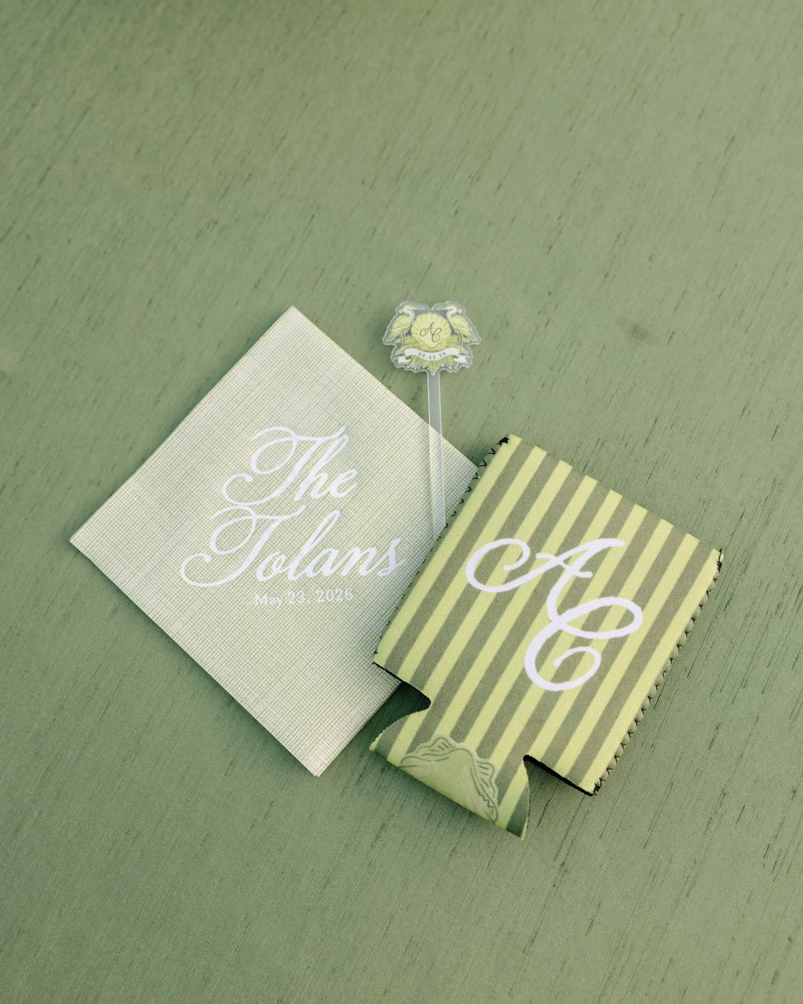 Green table with a white and gold wedding program, a striped folder, and a small sign with wedding initials and date.