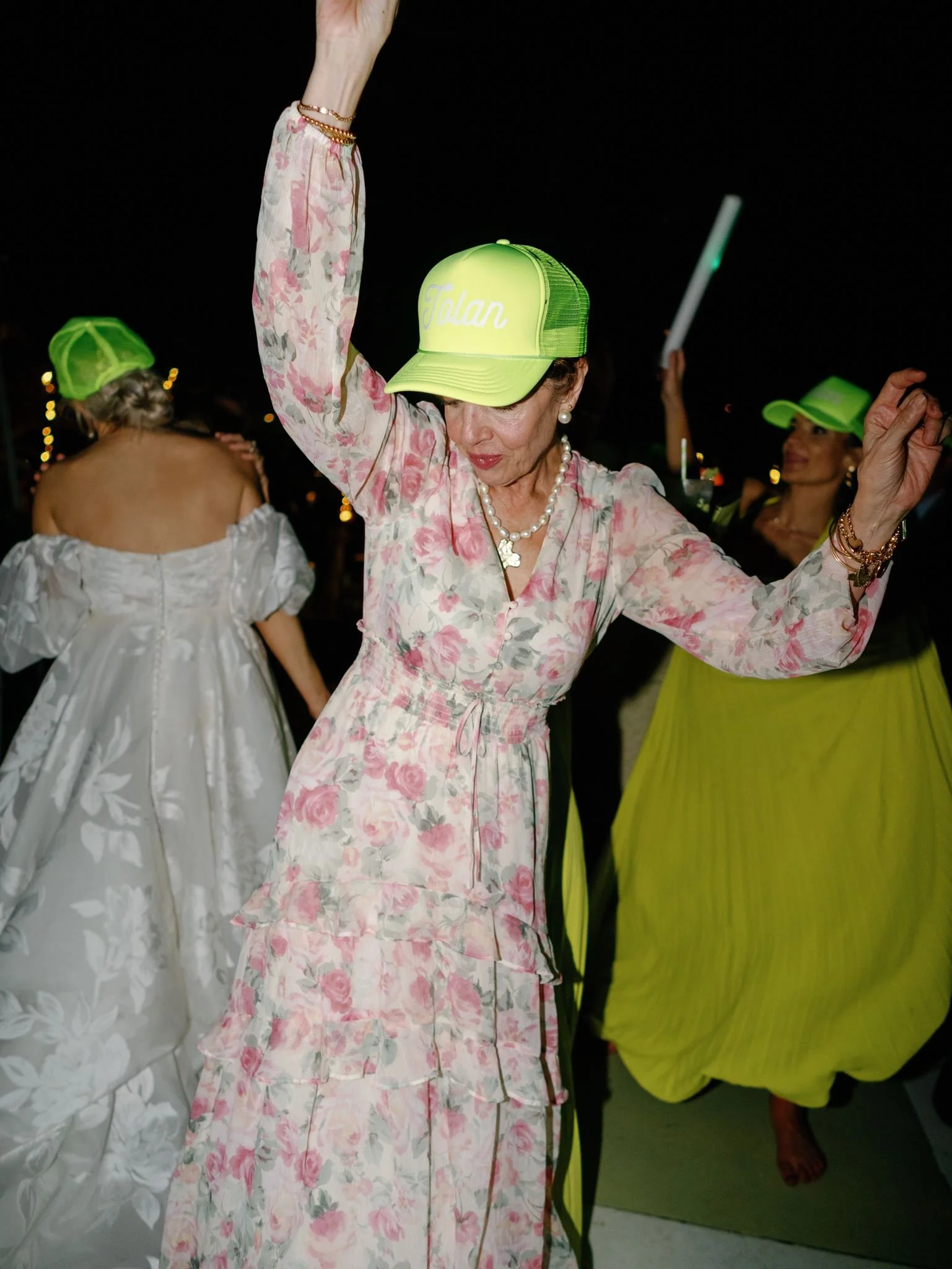 An elderly woman dancing at a nighttime event, wearing a pastel floral dress, pearl jewelry, and a neon yellow cap with the name "Tolan" embroidered on it.