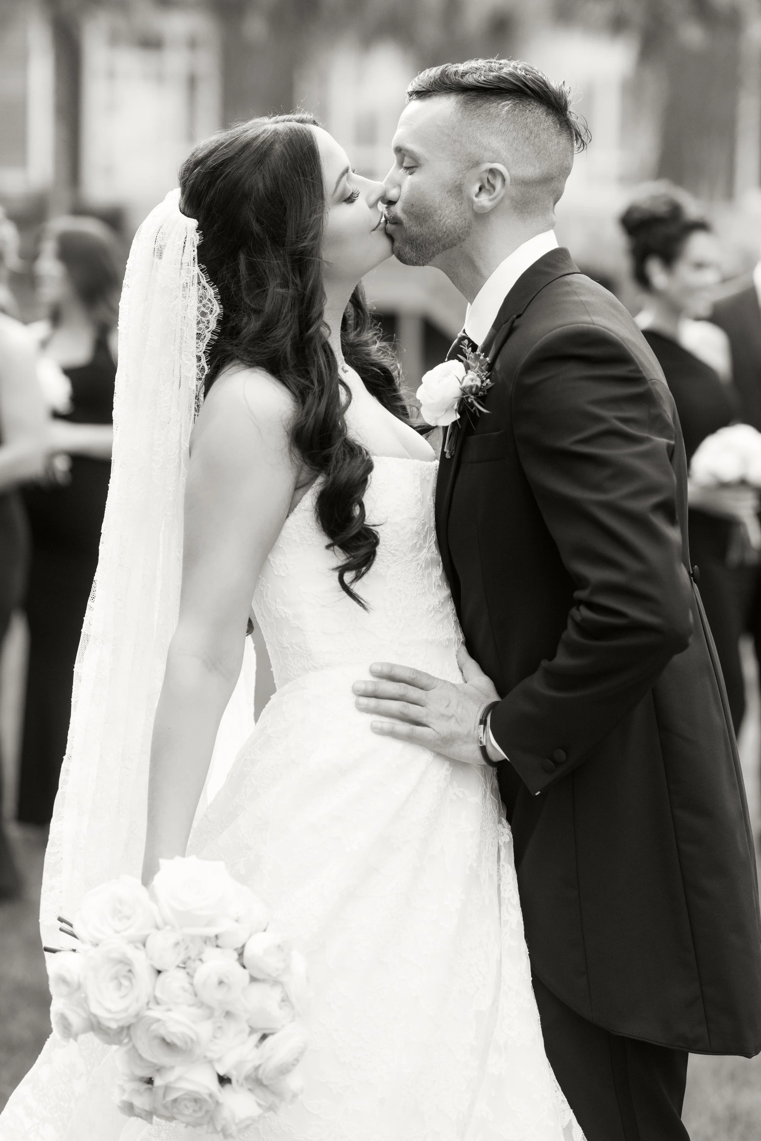 A bride and groom sharing a kiss during their wedding ceremony. The bride has long dark hair, wears a lace wedding dress and a veil, and holds a bouquet of roses. The groom has short dark hair, a beard, and is dressed in a dark suit with a boutonnièr