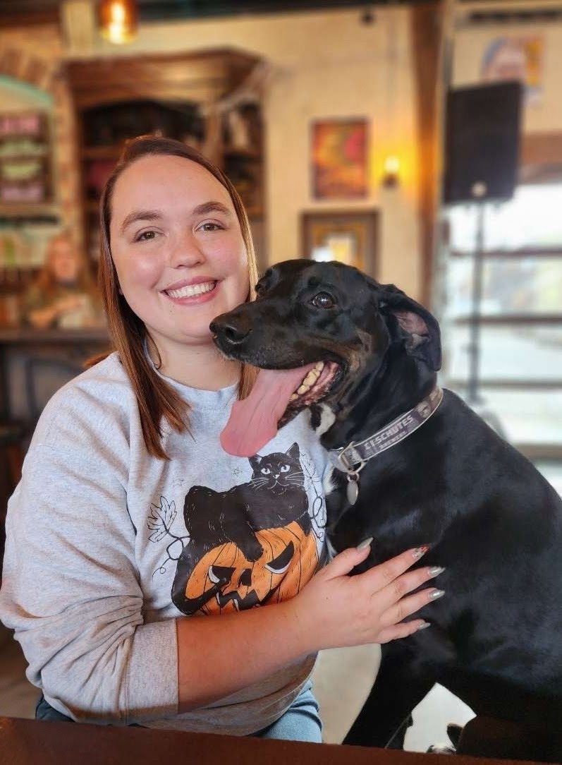 A smiling woman sitting inside a cozy café or restaurant, holding a large black dog with its tongue hanging out.