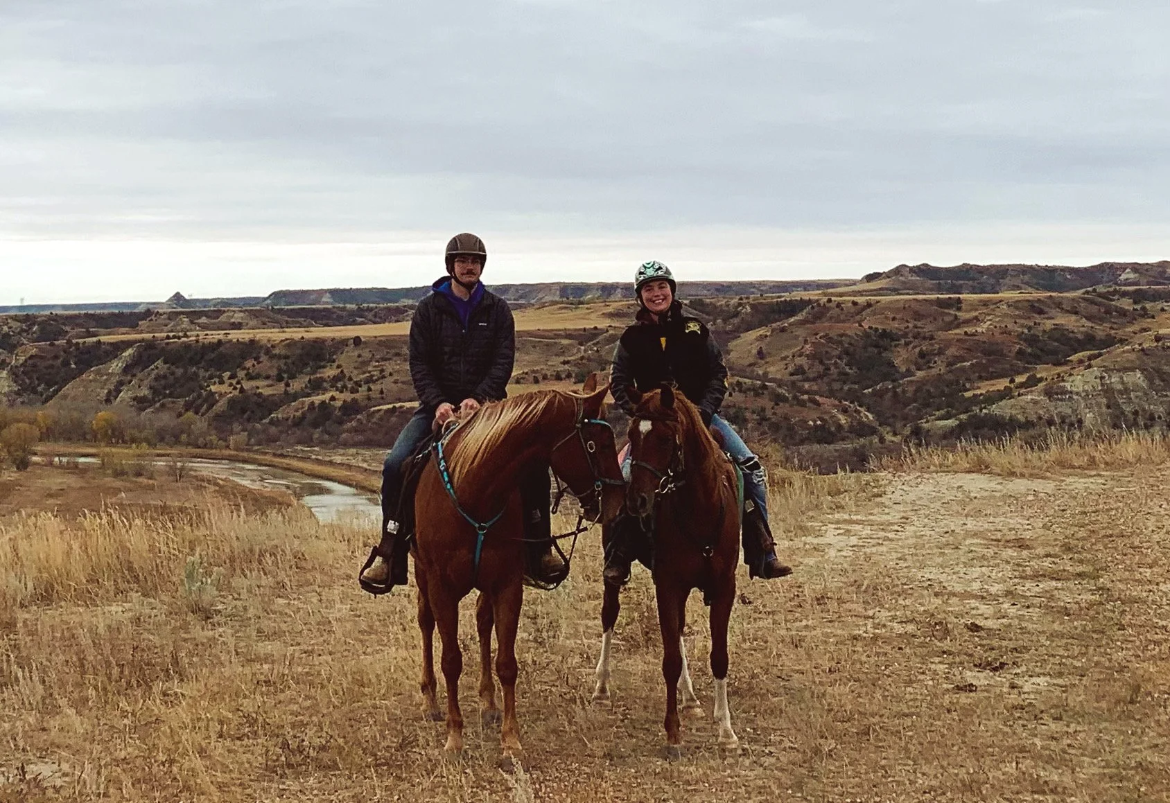 Two people riding horses in a scenic, hilly landscape under a cloudy sky.
