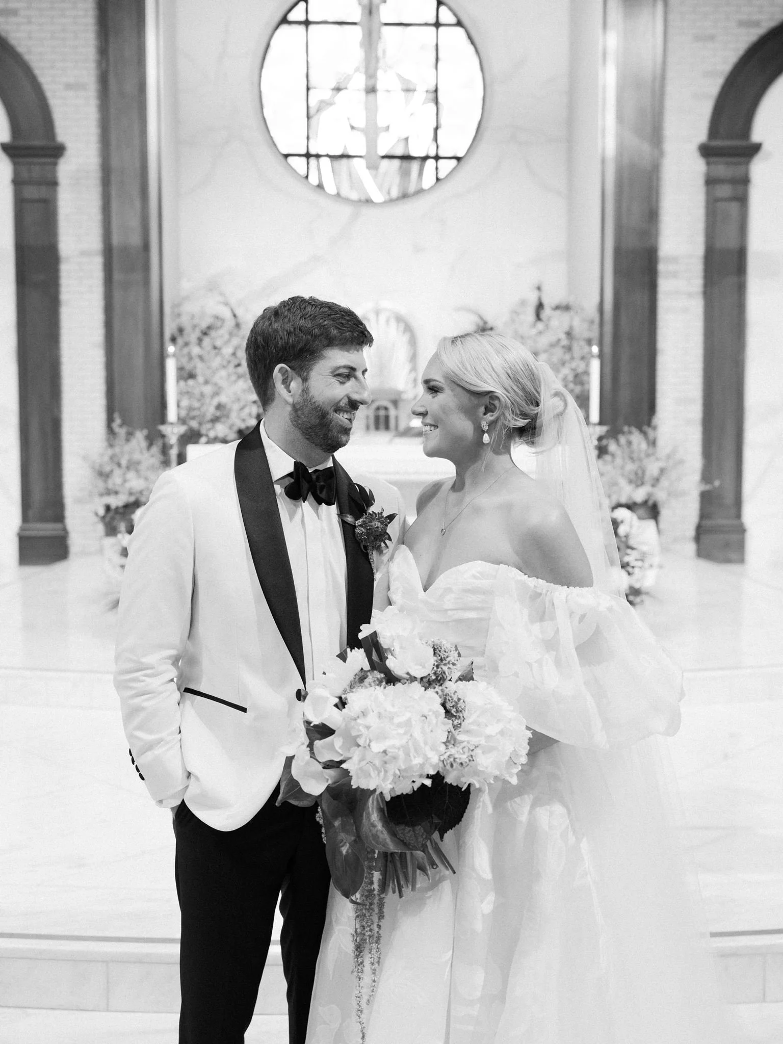 A newlywed couple in wedding attire standing inside a church, sharing a joyful moment. The groom is wearing a tuxedo with a bow tie, and the bride is holding a bouquet of flowers, wearing a wedding dress, earrings, and a veil. They are smiling and lo
