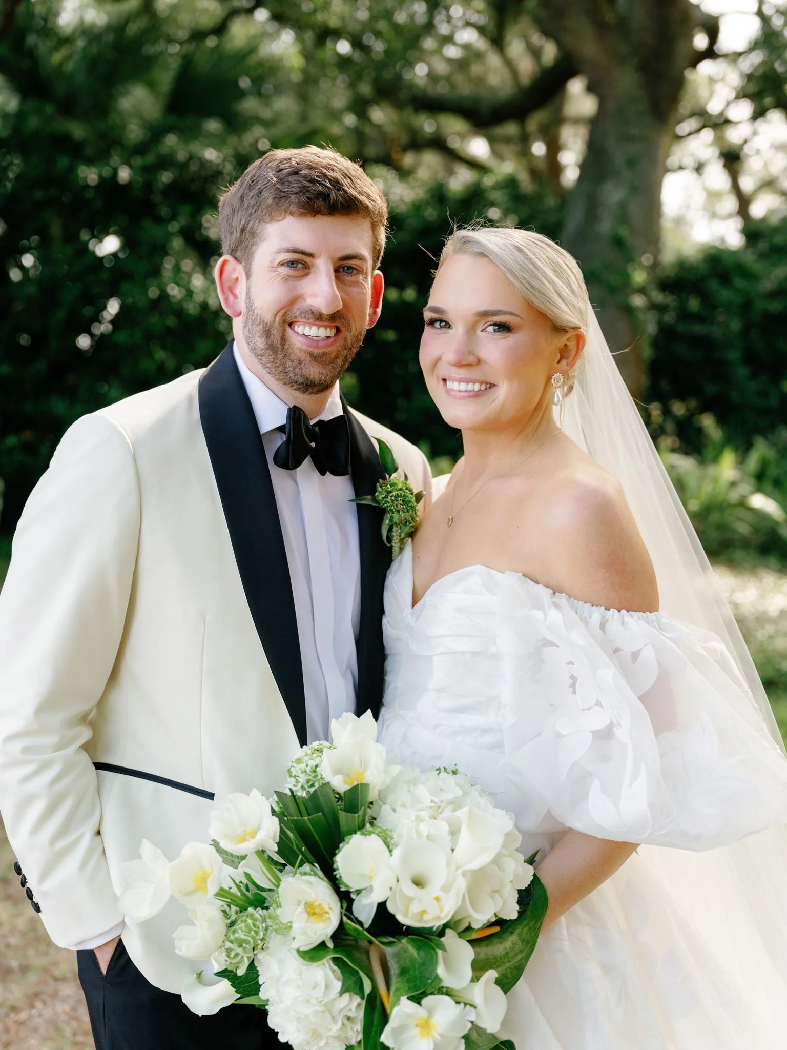 A smiling bride and groom dressed in wedding attire outdoors with trees in the background, holding a bouquet of white and yellow flowers.