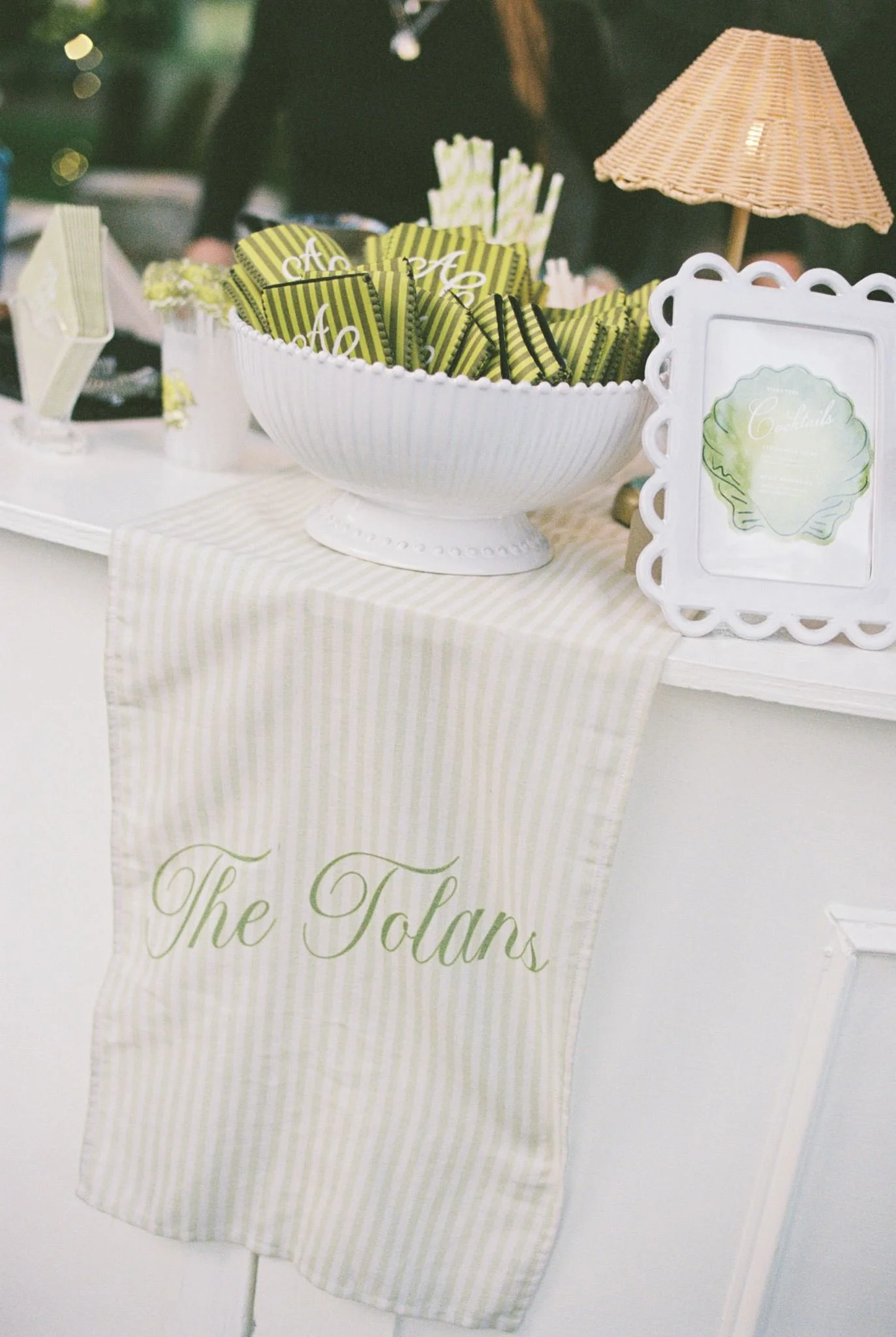 A table with a white tablecloth and a linen runner, featuring a large white bowl filled with green-and-black striped favor bags, a framed decorative sign with the word "Cocktails," a small umbrella, and small floral arrangements in vases.