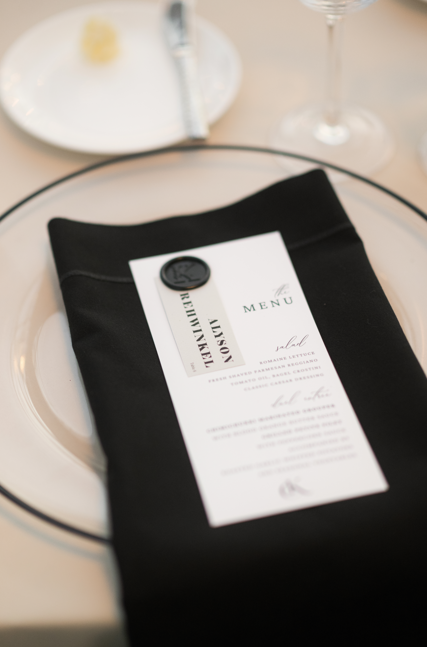 Restaurant table setting with a black napkin holding a printed menu, a small wax seal on the menu, a white plate, a butter pat, and a wine glass in the background.
