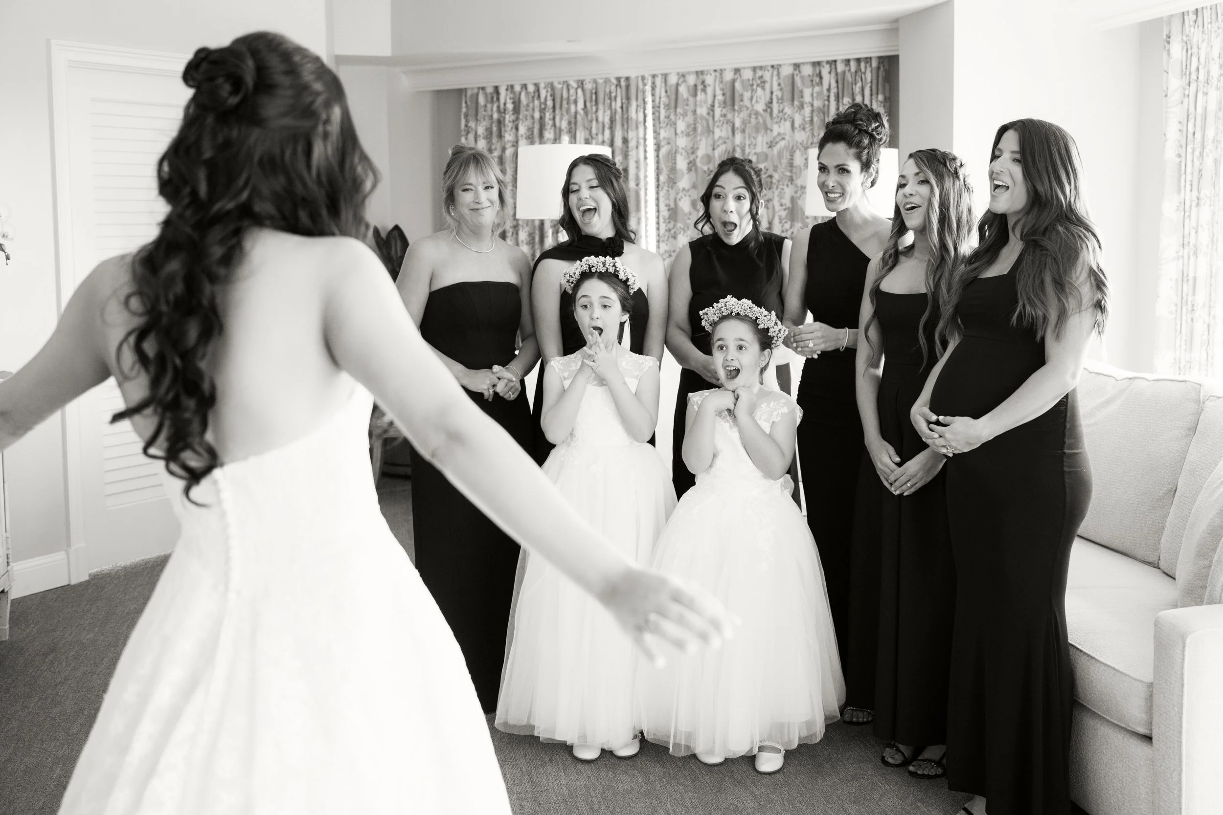 A woman in a wedding dress facing a group of women and two young girls, who are smiling and reacting with surprise and joy.