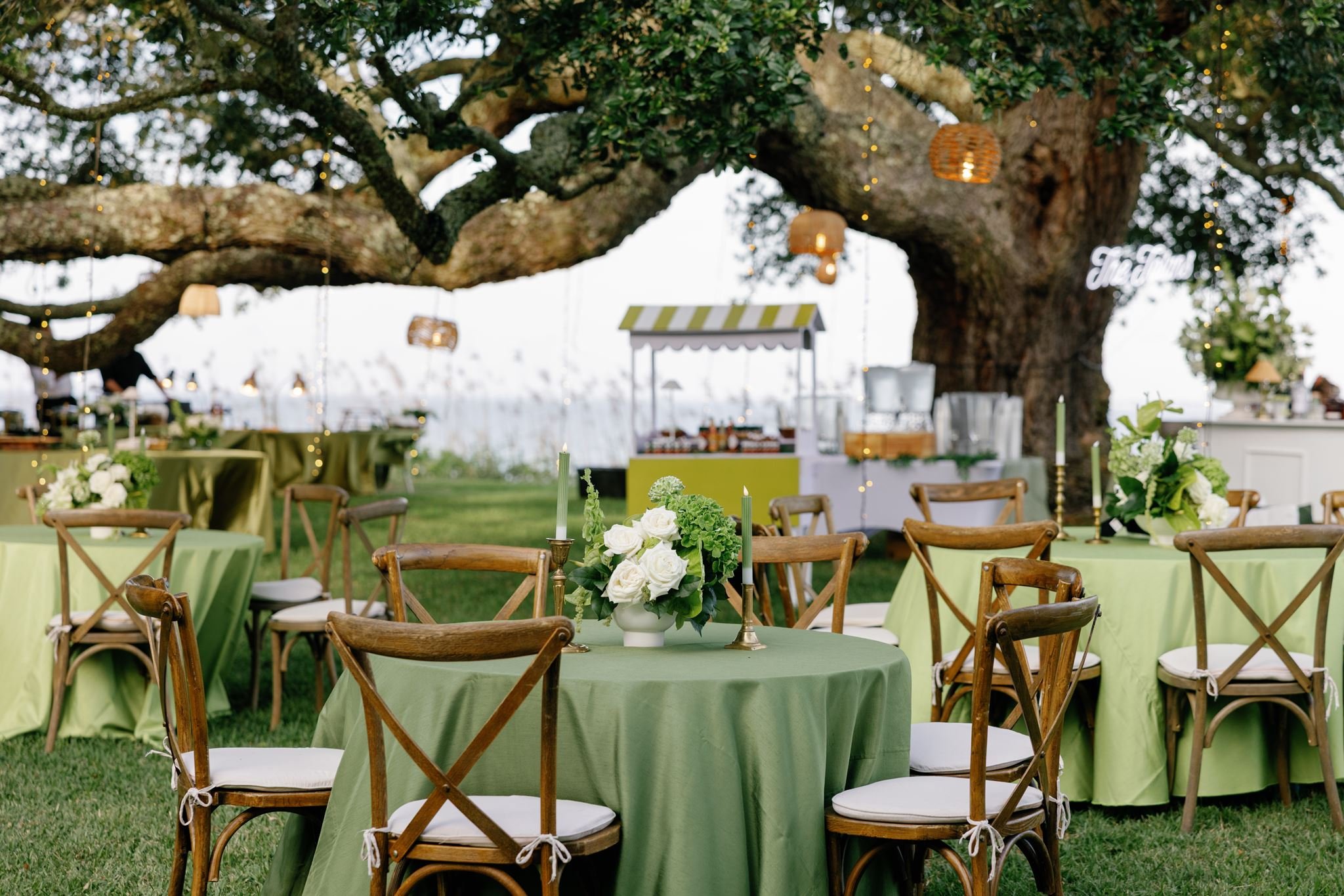 Outdoor event setup with round tables covered in light green tablecloths, wooden chairs with white cushions, floral centerpieces, and a large tree decorated with hanging lanterns and string lights.