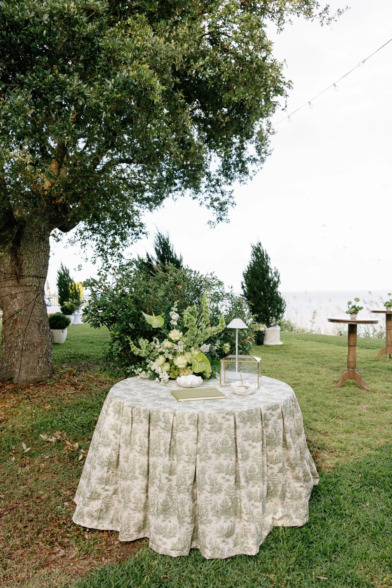 Outdoor event setup with a round table covered in a patterned tablecloth, decorated with a floral arrangement, lanterns, and a book, under a large tree with string lights.