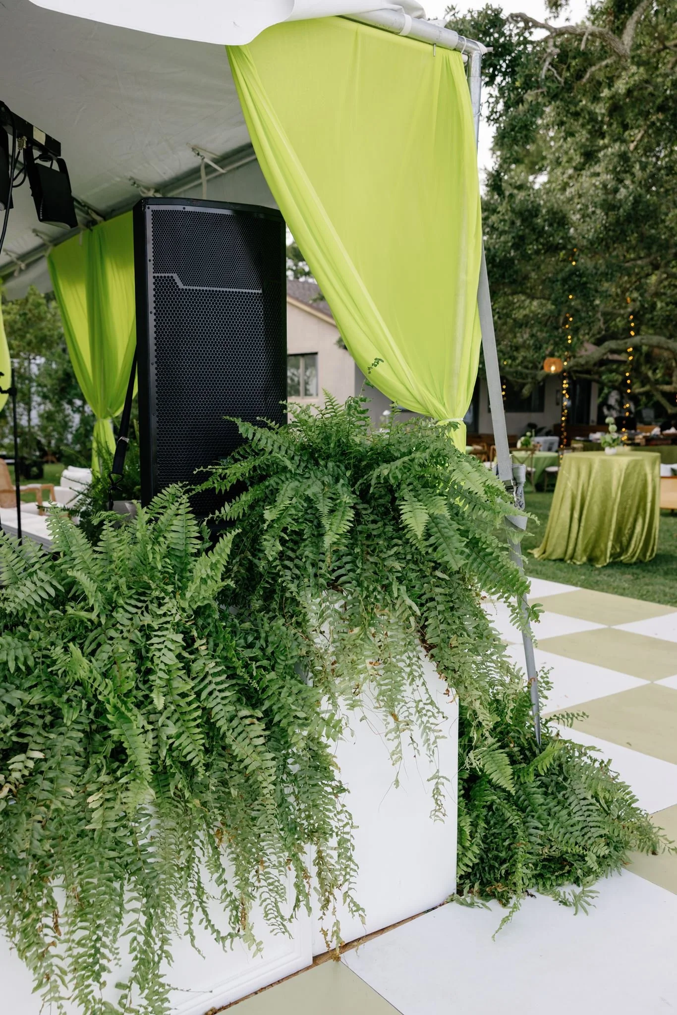 Outdoor event setup with green curtains, large black speaker, greenery, tables with green tablecloths, and string lights.