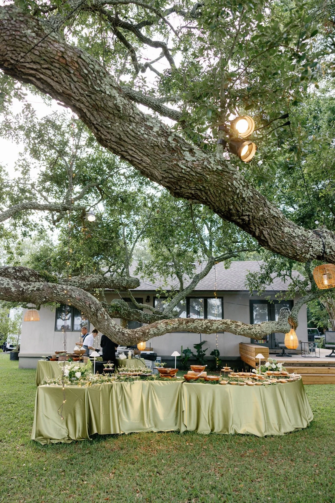 Outdoor wedding reception under large tree with hanging lanterns, featuring a table with yellow tablecloth and food setup, with people in the background near a house.